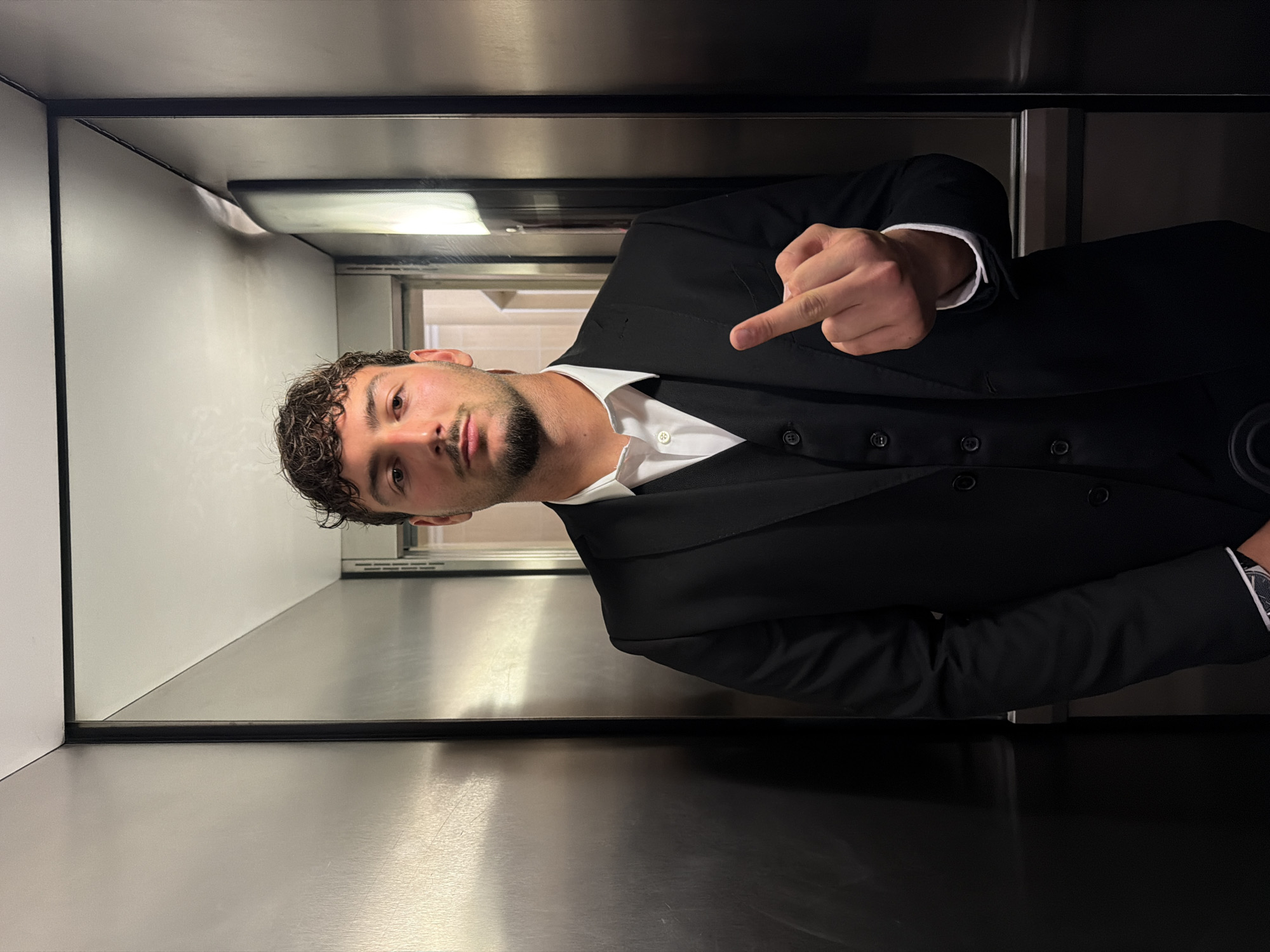 A man is standing inside an elevator cabin. He is wearing a black suit with a white shirt and giving the middle finger to the camera. The interior of the elevator has metal walls. There is a window visible in the background. The photo appears to have been taken inside the elevator of Granpeixe Shopping Vila Real, in Vila Real, Portugal.