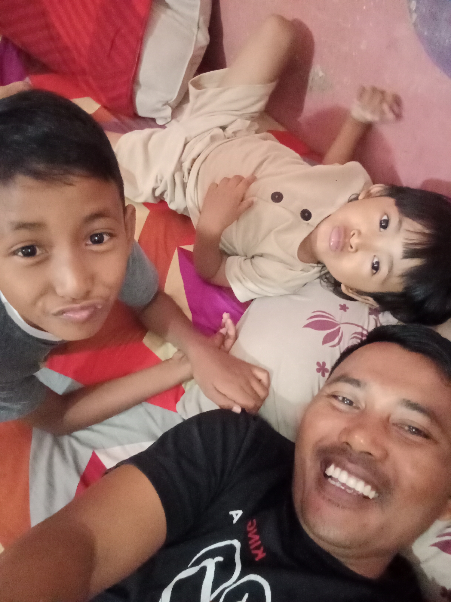 The image, taken indoors in Bloro, Indonesia, shows three individuals lying on a bed with a patterned bedspread. A man in the foreground, wearing a black t-shirt with white lettering that reads "KIME", smiles broadly, revealing his teeth. To his left, a young boy with dark hair and wearing a grey t-shirt looks towards the camera with a playful pout. Between the man and the boy, a young child, possibly a girl with dark hair, lies on her back, looking up. She is wearing a light-colored t-shirt with two dark buttons. The background consists of a red and pink patterned blanket and a glimpse of a white pillow. The lighting suggests an indoor setting, possibly a bedroom.
