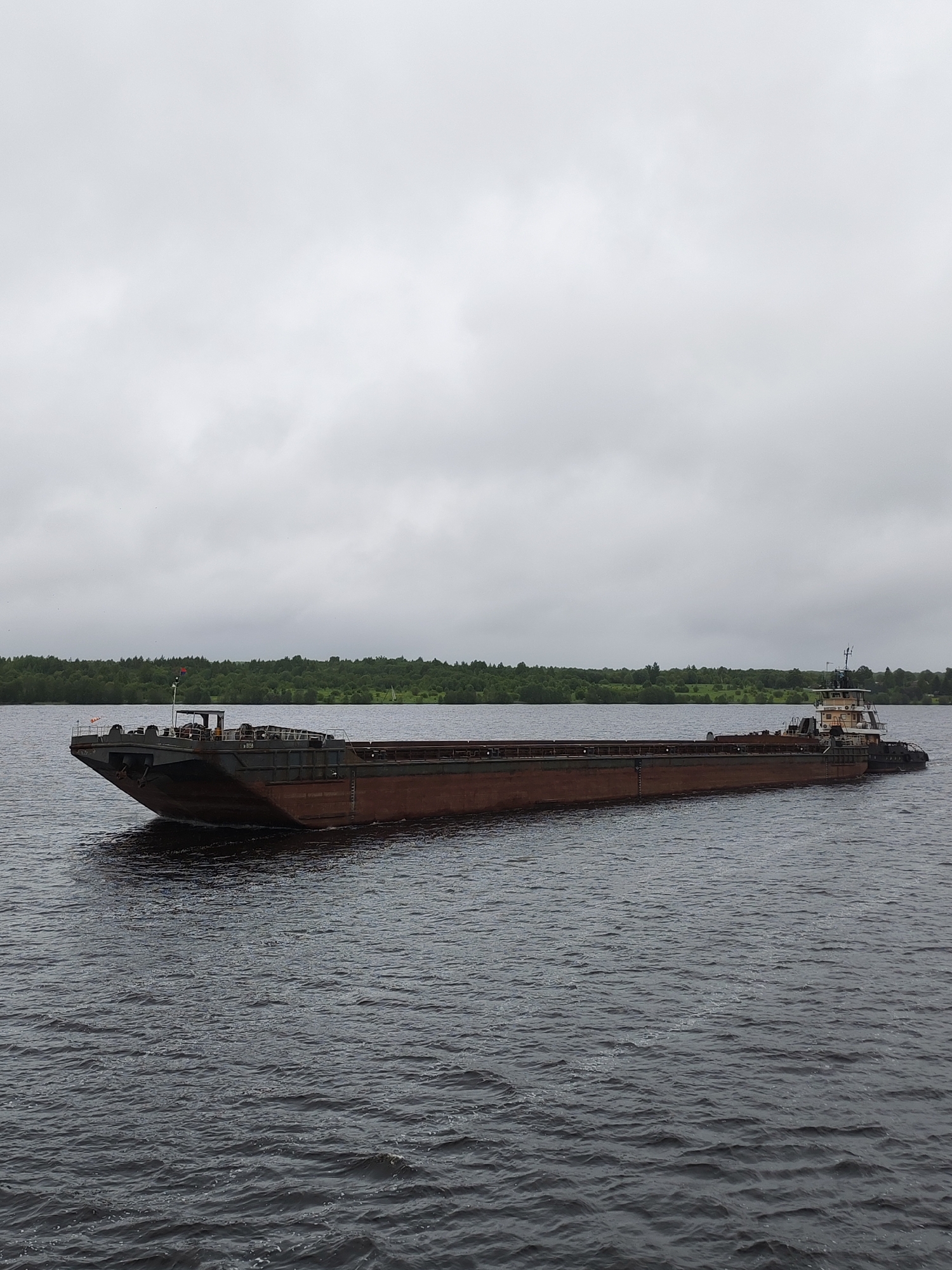 A barge is on the water in Раменье, Russia. The sky is overcast. The water is dark and slightly rippled. A thin strip of green trees lines the horizon.