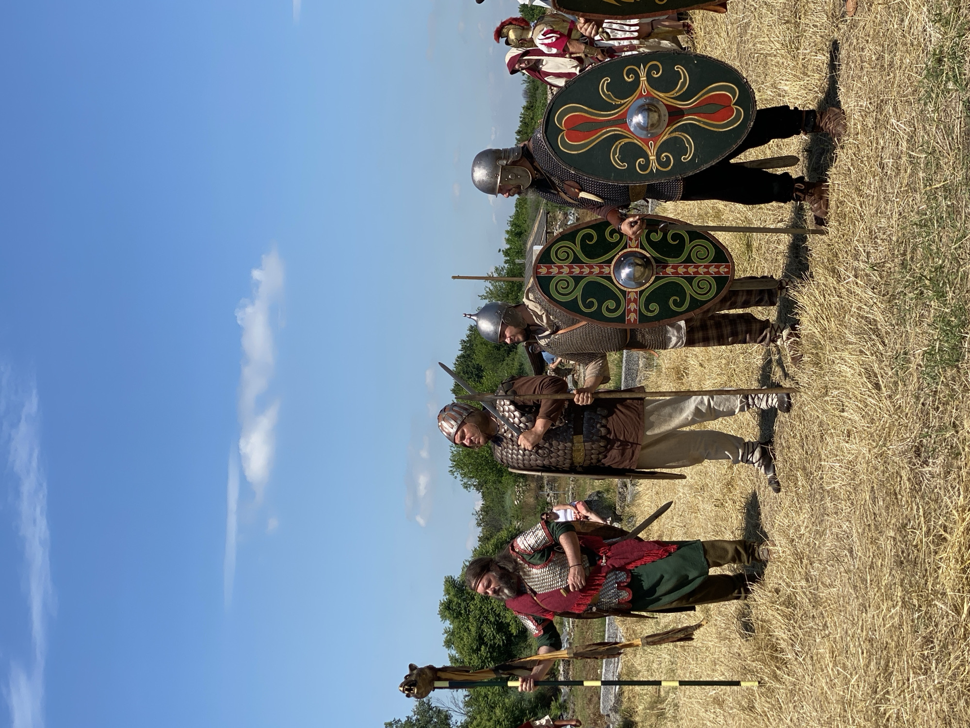 This image captures a historical re-enactment event in Gigen, Bulgaria, under a clear blue sky with scattered white cumulus clouds. Five individuals, dressed in period-specific military attire, stand in a line on a field of dry, yellowed grass, illuminated by bright daylight.
From left to right:
1. A man with a long dark beard and hair, wearing a green tunic, a red cloak or sash, and scale armor. He holds a tall, dark staff topped with a golden, bear-like animal head, consistent with a Draco standard. A sword is visible at his left hip.
2. A man with a beard and mustache, wearing a brown tunic, lamellar armor, light-colored trousers, and strapped footwear. He wears a distinctive ridged, conical metal helmet and holds a spear vertically.
3. A man with a beard, wearing a chainmail-like garment visible at his neck and sleeves, plaid trousers, and a metal helmet. He carries a dark green round shield adorned with red and gold Celtic-style spiral patterns and a prominent central metal boss. He also holds a spear.
4. A man with a beard and mustache, wearing dark clothing with visible chainmail, and a metal helmet. He holds a dark green round shield featuring intricate gold and red swirling motifs and a central metal boss. A spear is held in his right hand.
5. Partially visible on the far right, a figure in a red-plumed helmet and red and white Roman-style military attire, with armored shoulders.
The background consists of dense green foliage, including trees and shrubs, with indistinct white structures or ruins visible further back. A few additional, indistinct figures are visible among the foliage in the mid-ground. The individuals are posed, looking generally forward or slightly upwards.