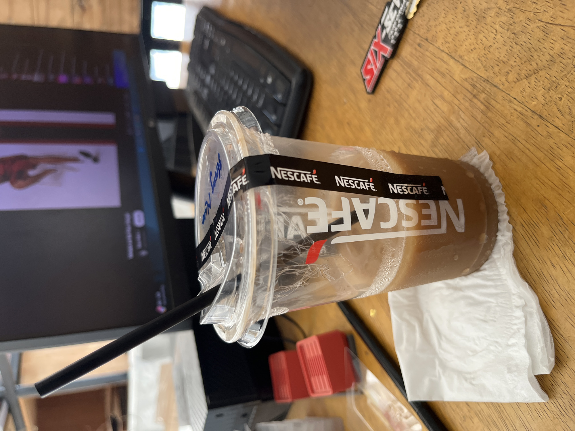 A close-up, eye-level, medium shot captures a disposable plastic cup filled with a brown beverage, likely iced coffee, resting on a white paper napkin on a wooden desk. A black straw extends from the cup at an angle. The cup is adorned with a black band featuring the "NESCAFÉ" logo repeated multiple times. In the background, a computer monitor displays an image, and a black keyboard is visible. To the left of the cup are two red, stacked objects, possibly small boxes. A black pen lies horizontally in the foreground to the left of the cup. A red and black sticker or keychain with the word "SIX" is positioned to the right of the cup, closer to the foreground. The lighting suggests an indoor environment, possibly an office or workspace. The scene implies a casual setting where someone is taking a break. The image does not provide specific clues to pinpoint the location in Kedawung, Indonesia.