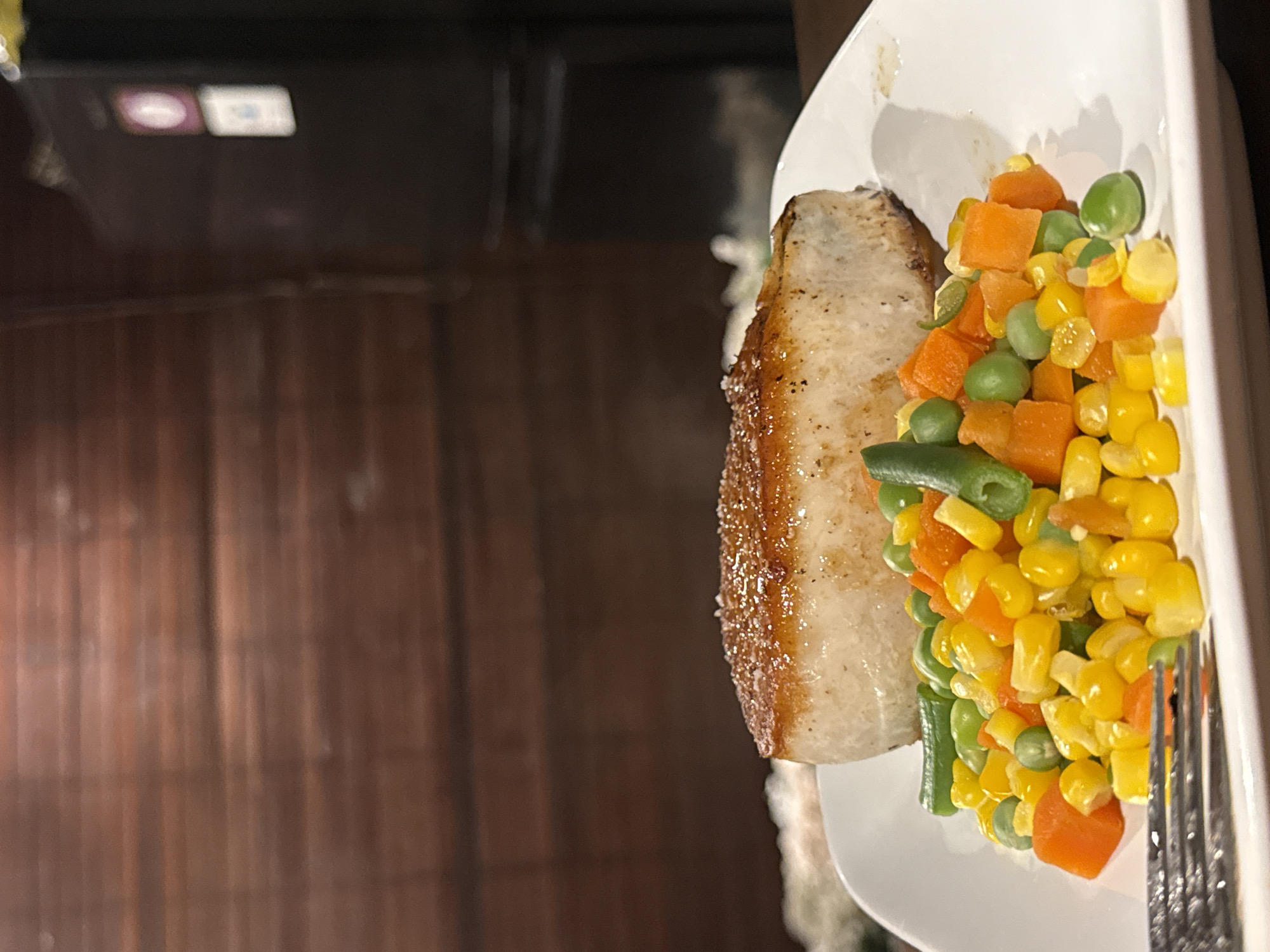 This media file shows a close-up of a plate of food, rotated so that the food is on the right side of the frame and the background is on the left. The plate contains a piece of white fish with a golden-brown crust, seasoned with black specks. To the right of the fish is a medley of mixed vegetables, including diced carrots, corn kernels, and green peas. A fork is visible on the right edge of the plate, partially submerged in the vegetables. The background on the left is blurred but appears to be a dark, textured surface, possibly wood or bamboo, with some indistinct shapes of possibly artificial lighting in the upper left corner. The lighting suggests it might be evening or indoor lighting. There is no indication of people or specific weather conditions. The scene is a close-up of a prepared meal, possibly at a restaurant or home.