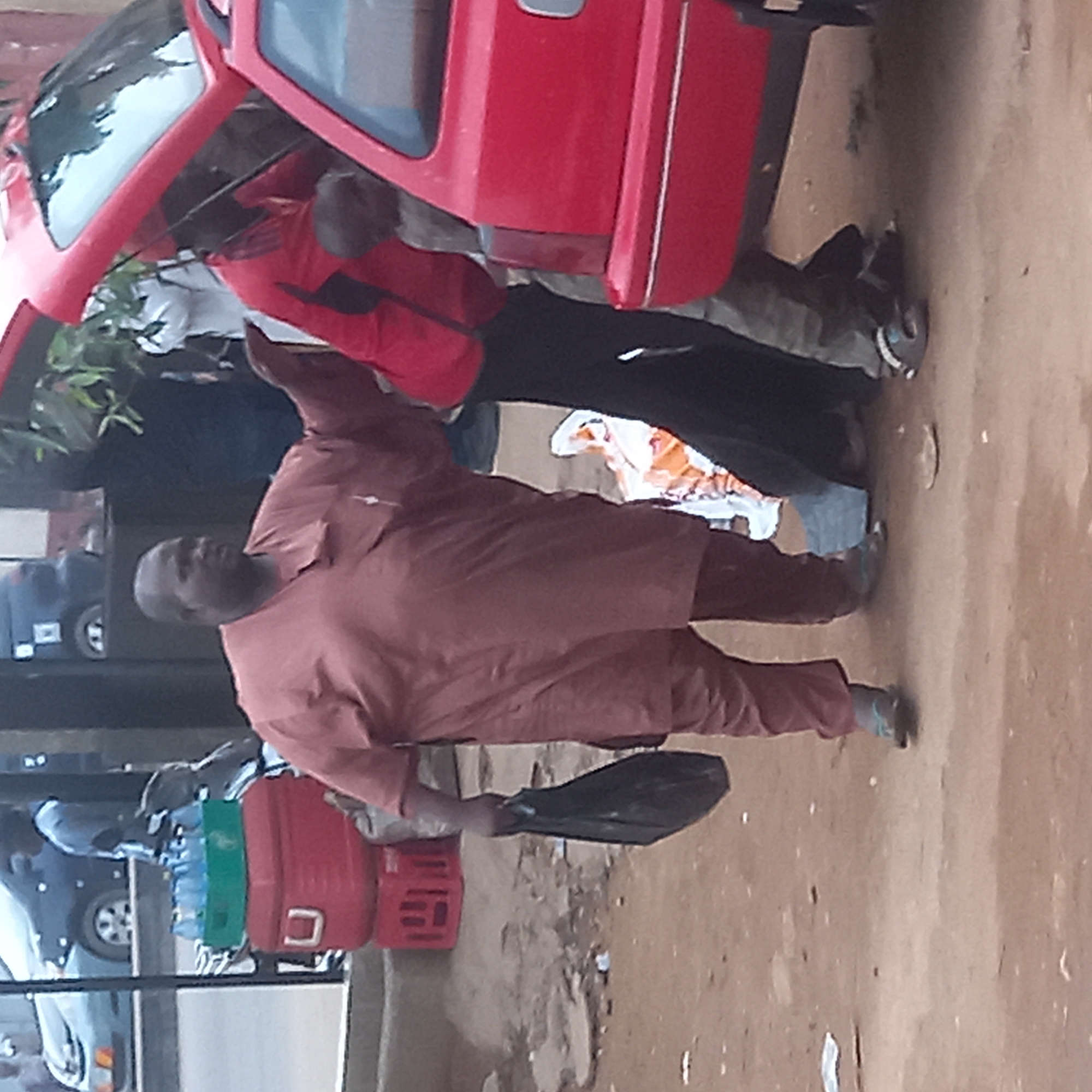 A person in a brown outfit stands on a dirt road in what appears to be an urban environment. They hold a black plastic bag in their left hand. A red car is visible to the left, with two people standing near its open hood. Another person is standing next to the car wearing a red jacket. Several red and green crates are stacked together nearby. There are other cars parked in the background. The location is Dadi Riba, Nigeria.