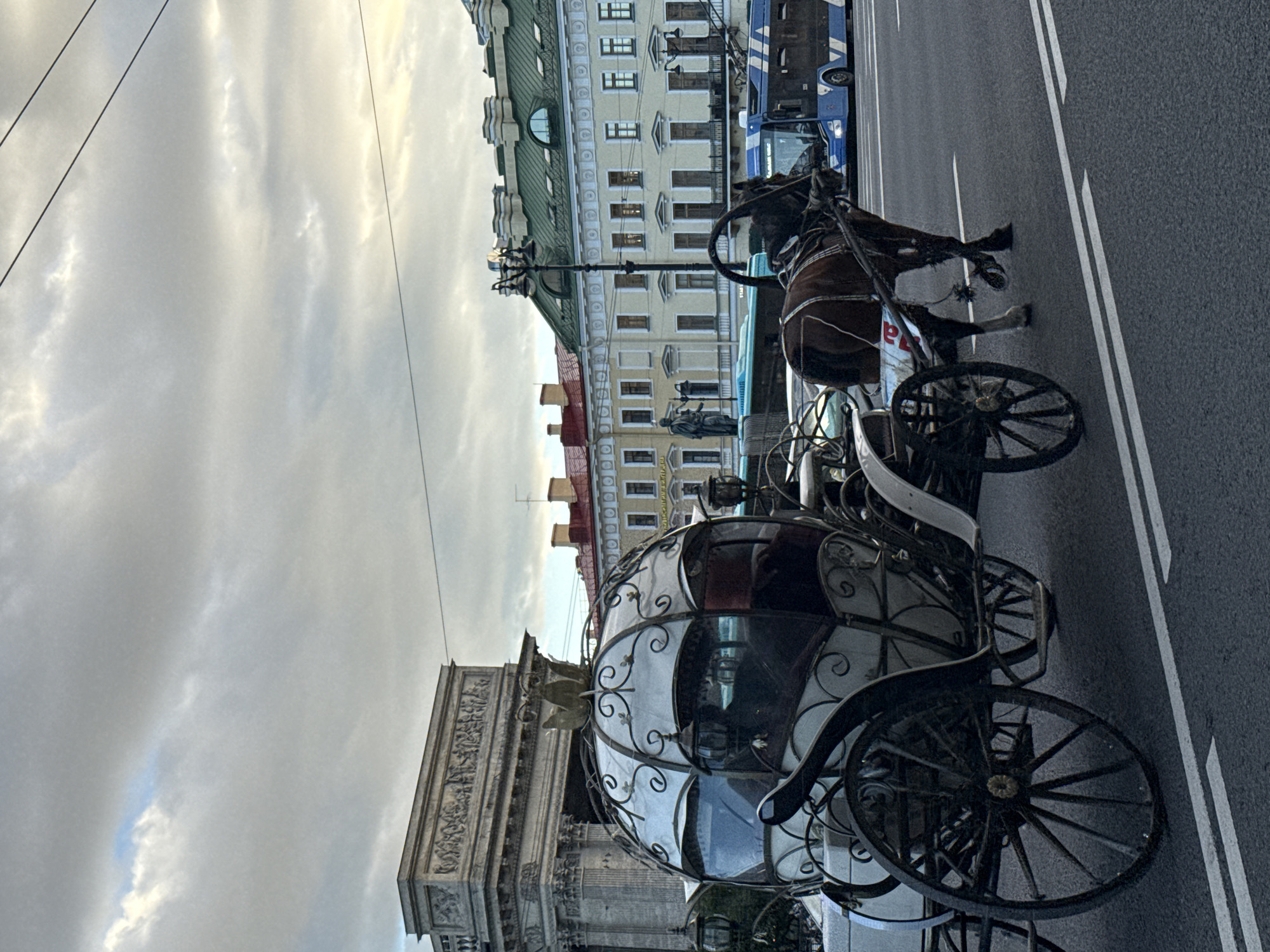 The image shows a horse-drawn carriage and a blue bus on a city street. The carriage has a white, ornate body with intricate metalwork and a golden crown-like embellishment on top. A brown horse is harnessed to the carriage, wearing a bridle and harness. In the background, there are several buildings with classical architecture, some featuring columns and ornate facades. One building has a green roof. A large, triumphal arch-like structure with detailed carvings is visible on the left side of the image. The sky is overcast with dramatic clouds. The street has white lane markings. The presence of the horse-drawn carriage suggests a tourist area, likely in the historic center of Saint Petersburg, Russia, given the architectural style and the presence of a monument or triumphal arch which is likely part of the historic cityscape.