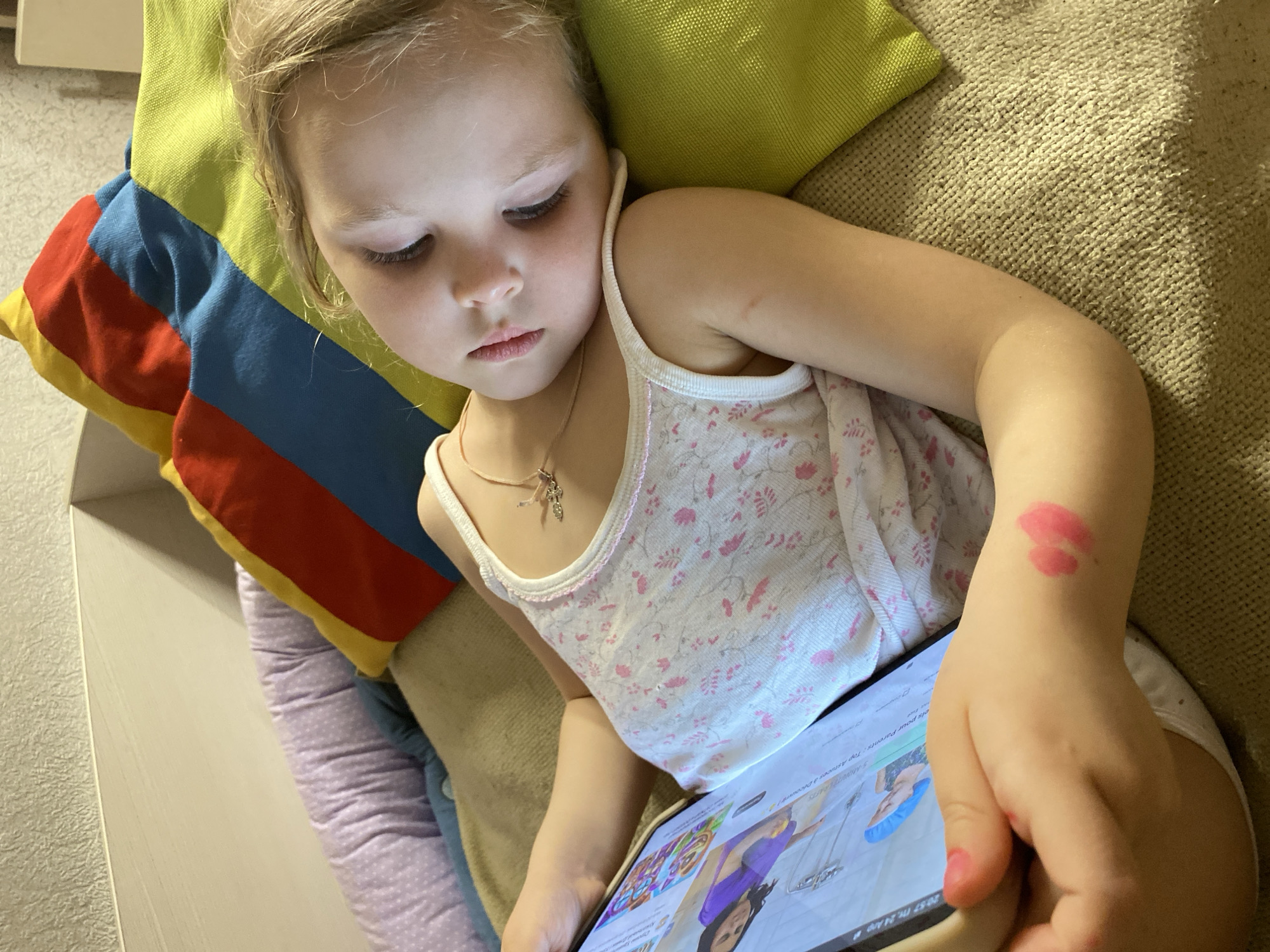 A young blonde child, likely a girl around preschool age, is captured in a close-up, lying down and engrossed in content on a tablet. She wears a white sleeveless top adorned with a subtle pink floral pattern and a delicate necklace with a small charm. Her eyes are focused intently downwards on the tablet screen, which casts a soft illumination on her face, revealing a calm and concentrated expression.
A distinctive red lipstick mark, shaped like a kiss, is visible on her left forearm, and one of her fingernails is painted with pink polish. She is surrounded by colorful cushions: a bright lime-green one behind her head and another with bold horizontal stripes of red, blue, and yellow. A light purple, possibly patterned, cushion is also partly visible. The background includes a textured light-colored wall on the left side of the frame.
The tablet screen prominently displays text that reads "5 Minute Crafts," suggesting the type of video or content being viewed. Below this, partially visible text reads "Parents: Top Astuces..." indicating a potential "tips for parents" theme. The device's status bar shows a timestamp "20:57," signifying that the scene is taking place in the evening. The child appears to be relaxing indoors, possibly on a couch or bed, in Votkinsk, Russia.