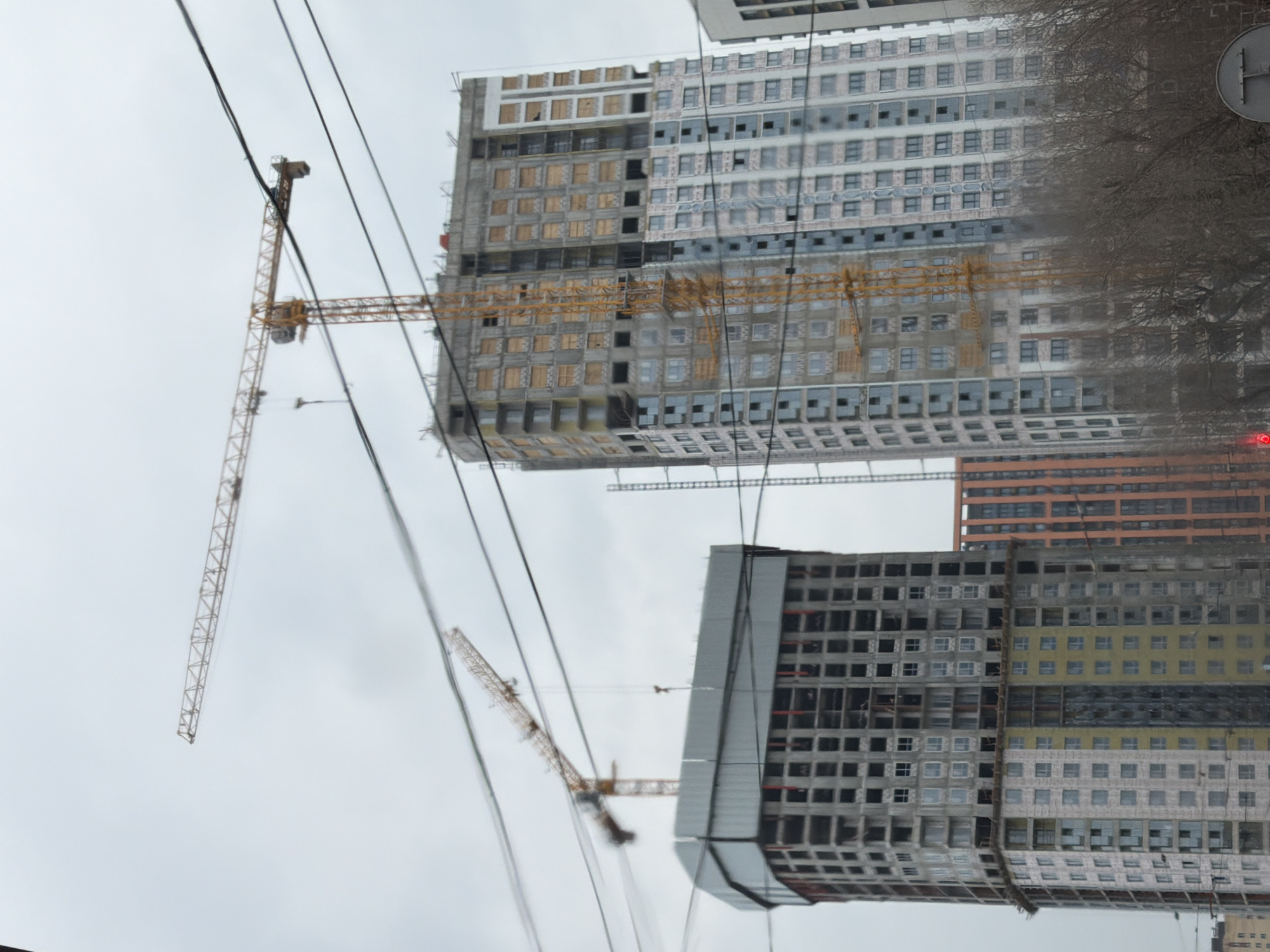 The image shows construction cranes and unfinished apartment buildings in Perm, Russia. The buildings have multiple floors and are in various stages of completion. The sky is overcast, and there are power lines in the foreground.