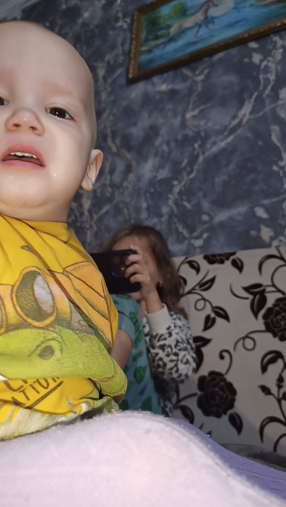 A close-up, low-angle shot focuses on the face of a bald infant in the foreground, with part of a yellow t-shirt visible. Behind the infant, a younger child holds a smartphone up to their face, appearing to take a photo. The background features a dark, marbled wallpaper and a patterned sofa. A framed painting of horses is visible on the wall above the sofa. The scene is likely indoors, possibly in a residential setting in the city of Karsha, Russia.