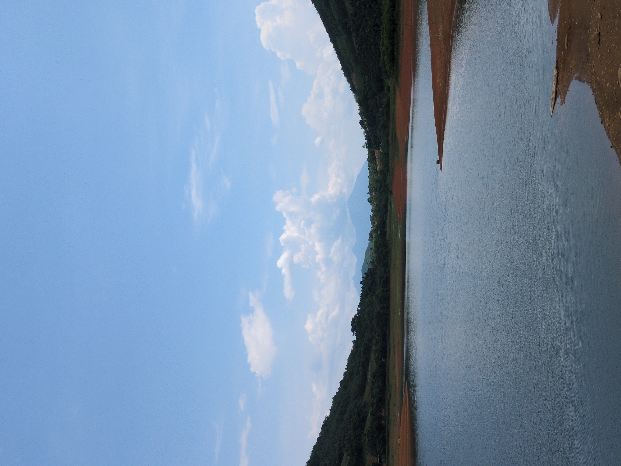 This image captures a serene natural landscape, likely a reservoir or lake, located in the vicinity of Ngọc Lặc Commune, Vietnam. The scene is dominated by a large body of calm, blue water that fills the foreground and midground. The water's surface exhibits gentle ripples, reflecting the pale blue sky above.
The shoreline is characterized by reddish-brown soil, particularly noticeable on the right side of the frame where it forms a gradual incline towards the water. Scattered on this exposed earth are a few small rocks and a piece of driftwood. Further inland, lush green hills rise steeply on either side of the water. These hills are densely covered with trees and vegetation, creating a natural backdrop. In the distance, hazy blue mountains are visible, adding depth to the landscape.
The sky above is a clear, bright blue, punctuated by scattered white clouds. Some of these clouds are small and wispy, while others are larger and more voluminous, particularly in the upper portion of the image. The overall impression is one of tranquility and natural beauty. There are no people or animals visible in the frame, and no man-made structures, aside from what appears to be a small dark object on a distant point of land, possibly a marker or a small structure, and the aforementioned driftwood. The lighting suggests a daytime scene, possibly late morning or early afternoon, with diffused sunlight.