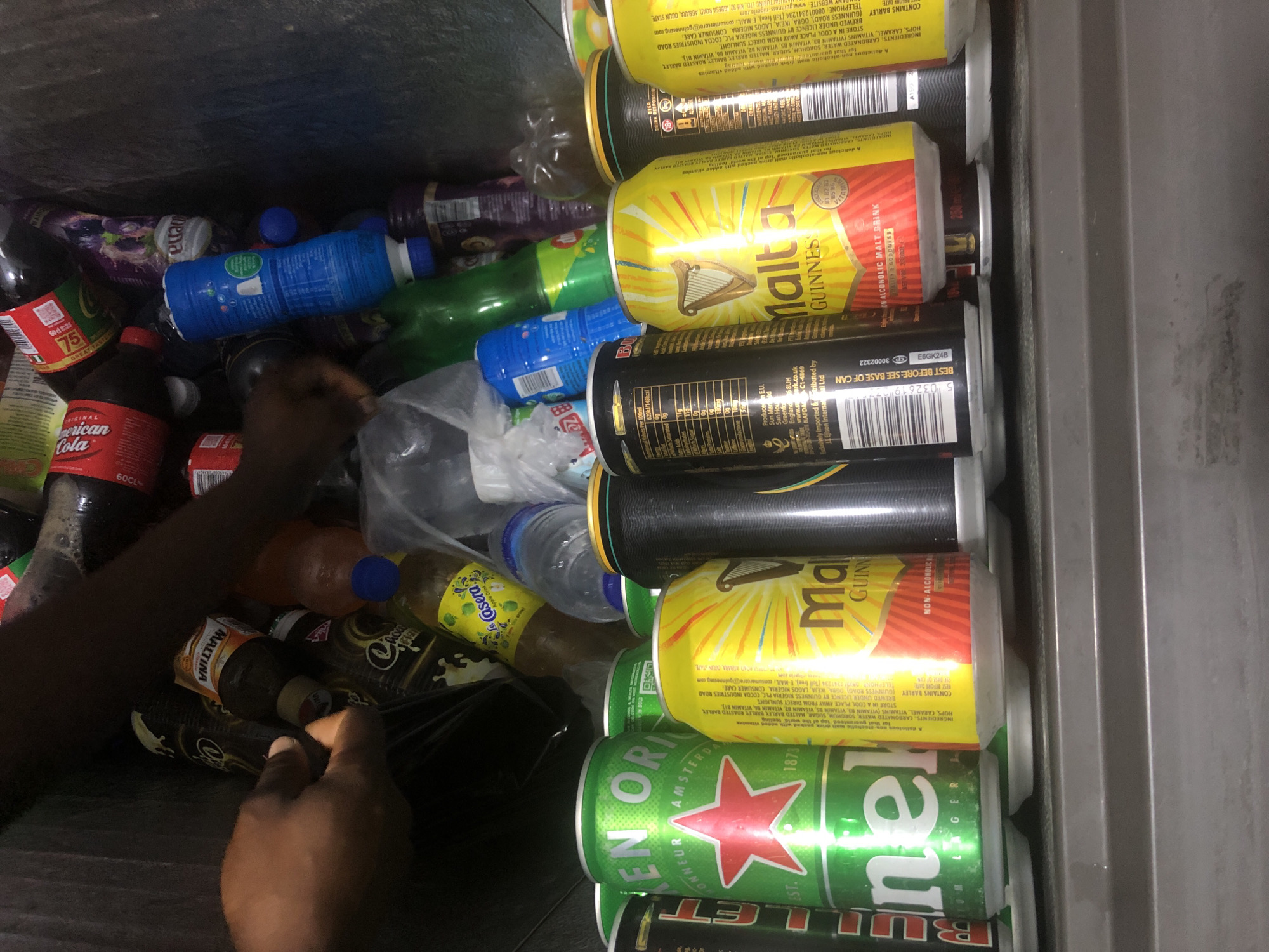 A man's hands reach into a refrigerator filled with various beverages, including cans of Heineken, Malta Guinness, and what appear to be bottles of Coca-Cola and other sodas. The refrigerator is packed tightly, with many drinks stacked on top of each other. The man's left hand is visible, touching a can, while his right hand is partially obscured by other bottles. The lighting is dim, suggesting the interior of a refrigerator or a poorly lit storage area. The overall scene depicts a person selecting or retrieving drinks from a chilled storage unit. The presence of specific brands like Heineken and Malta Guinness, along with the context of such beverages, suggests a location where these are commonly sold or consumed. Given the brands and the context, this scene could plausibly be in a commercial establishment such as a bar, restaurant, or a store in Ado, Nigeria, where these beverages are popular.
