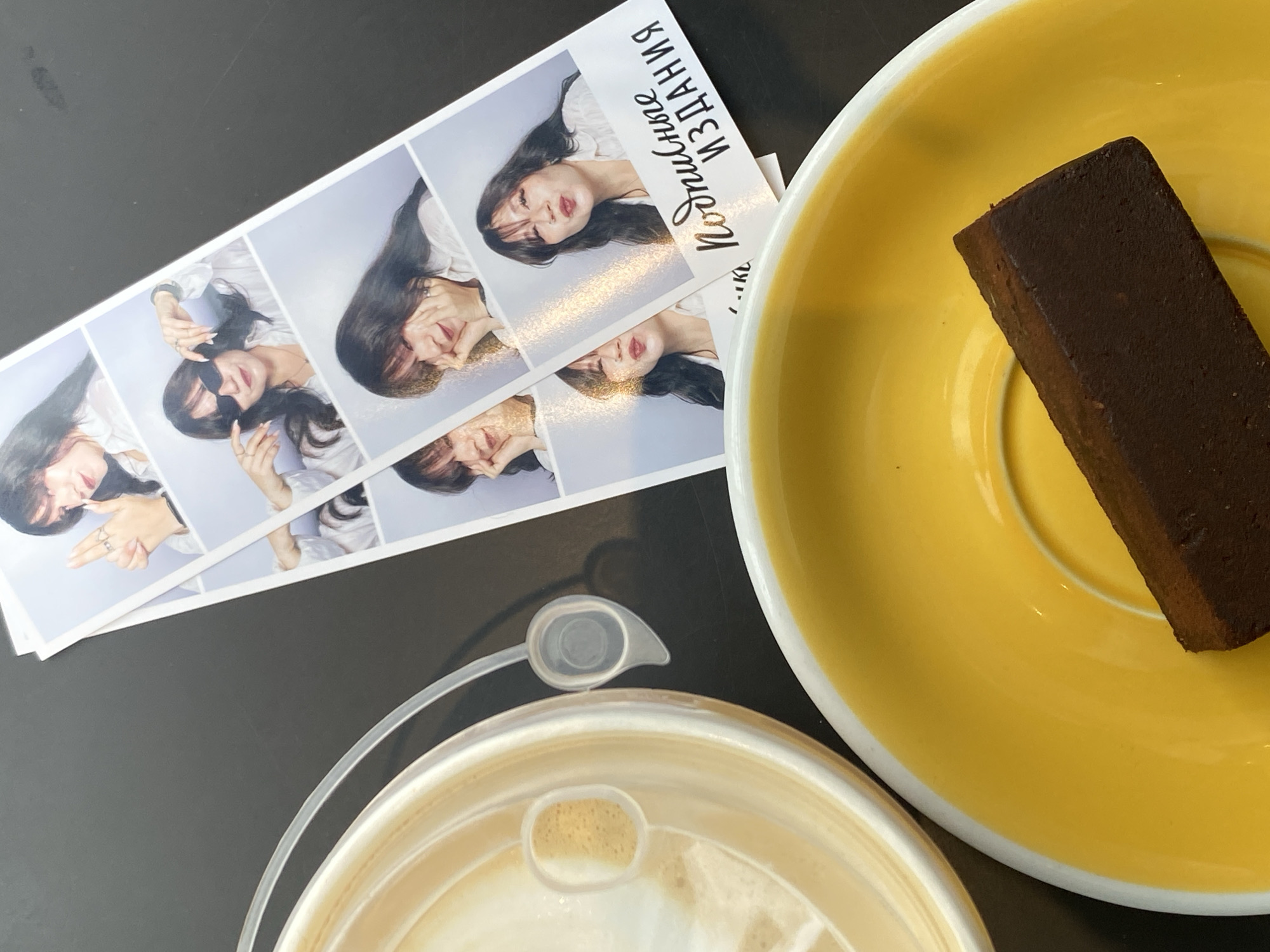An overhead shot depicts a dark, textured surface that appears to be a table. On the right side of the frame, a strip of photo booth prints shows a young woman with dark hair posing in four different frames. In the top frame, she is making a "finger gun" gesture with her right hand. The second frame features her wearing sunglasses and adjusting them. The third and fourth frames show her with her hands near her face, looking coyly at the camera. The bottom of the photo strip displays Russian text that reads "Подписные Издания" which translates to "Subscription Publications." To the left of the photo strip, a beige disposable coffee cup lid with a straw opening is visible. Partially beneath the lid and to the bottom right of the frame is a bright yellow saucer. On the saucer rests a rectangular piece of dark chocolate. The overall scene suggests a casual outing in a cafe or a similar establishment. The "Подписные Издания" text on the photo strip is a strong indicator that this scene took place in the city of Saint Petersburg, Russia, as this is the name of a well-known bookstore chain in the city.
