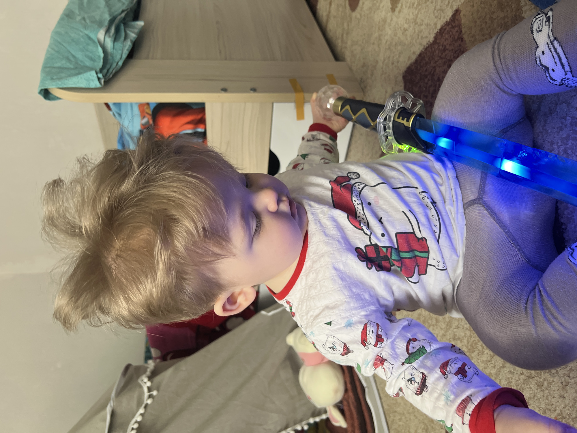 This image, taken in Votkinsk, Russia, features a young child, likely a toddler, positioned indoors, possibly on a bed or rug. The child has light blonde hair and is wearing white long-sleeved pajamas with red trim at the collar and cuffs. The pajamas are adorned with a repeating pattern of Christmas-themed polar bears, some wearing Santa hats and others holding wrapped presents. The child also wears grey tights, with a cartoon character (possibly a reindeer or snowman) visible on the right leg.
The child is holding a toy sword that emits a vibrant blue light from its blade. The hilt of the sword is dark-colored with gold accents and clear plastic elements. The child's eyes are downcast, indicating focus on the toy or a calm, contemplative state.
The background shows a light-colored wooden furniture item, possibly a bed frame or shelf, with a teal fabric draped over it. A light-colored plush toy, resembling a bunny or lamb, is partially visible near the child's left side. The child's lower body rests on a patterned rug with shades of brown and beige. The artificial lighting, combined with the glow of the toy, suggests the time of day is evening or night. No discernible text is visible in the image.