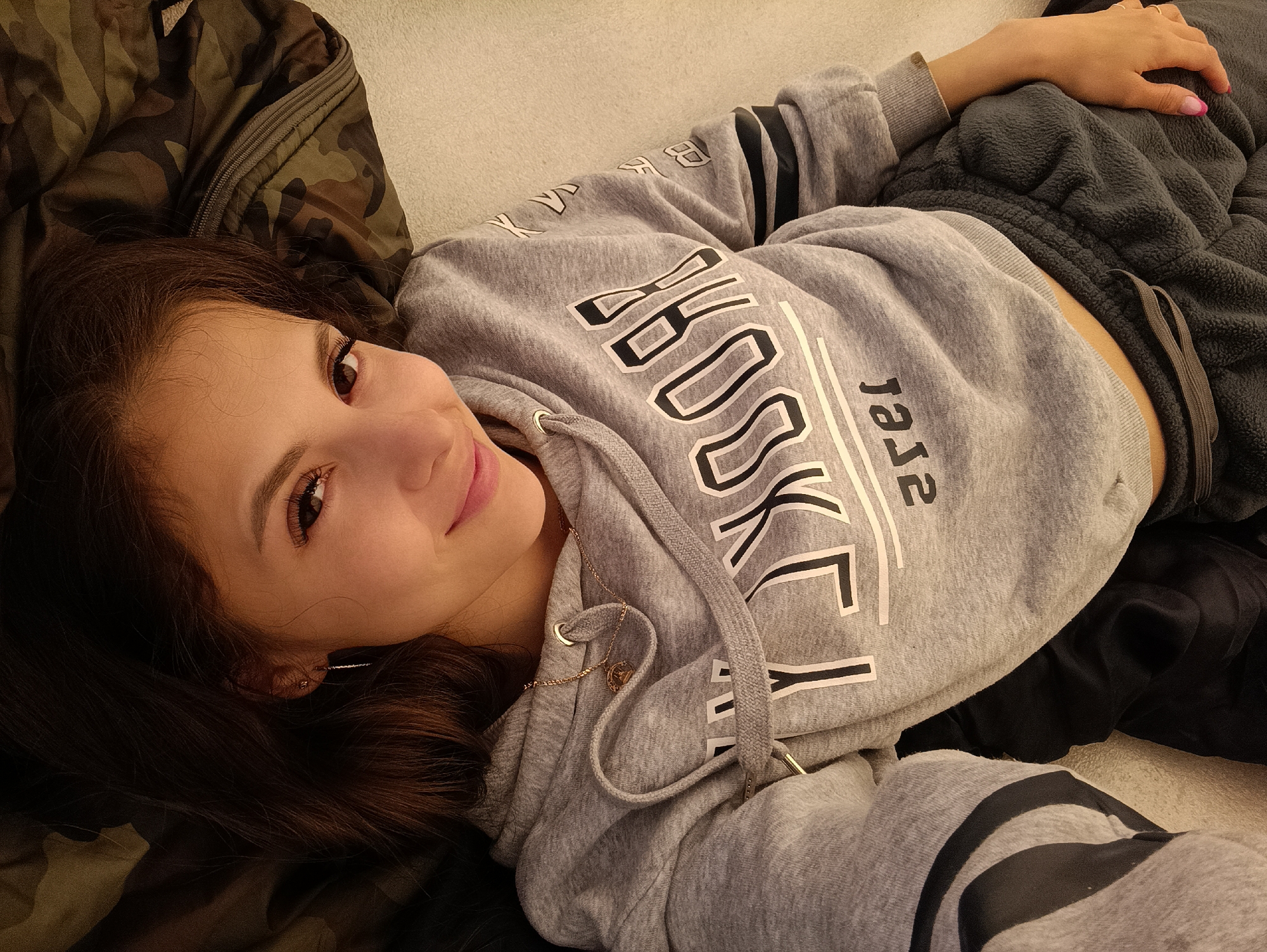 The image shows a young woman lying on her back. She has long, dark brown hair, and she is wearing a grey hooded sweatshirt with black stripes on the sleeves and "BROOKLYN" printed in large white letters across the chest. She is also wearing dark grey sweatpants. Her left hand is resting on her stomach, and she is wearing a ring with a red stone. The background consists of a camouflage jacket and a light-colored surface, possibly a bed or couch. The lighting appears to be indoor, with a soft glow. The woman has a slight smile and her eyes are looking towards the camera, suggesting a casual and relaxed pose. The overall mood is peaceful and intimate. The time of day and weather are not discernible from the image. No specific location cues are visible within the image itself to confirm the context of Sergeevka, Russia, beyond the provided prompt. The text visible on her hoodie is "BROOKLYN".