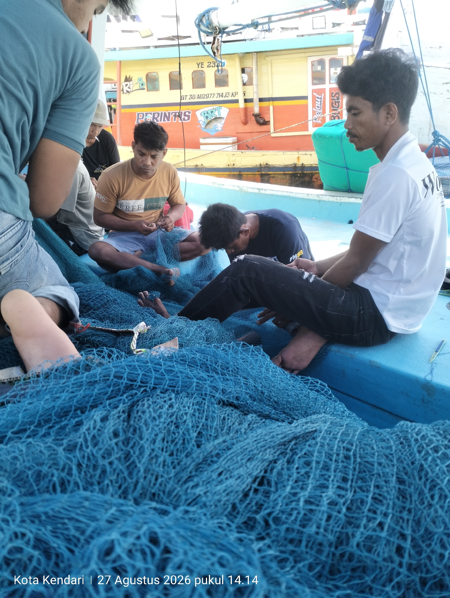 A group of four individuals are on a boat in the city of Kendari, Indonesia, on August 27, 2026, at 2:14 PM. They are surrounded by a large amount of blue fishing net. One person, on the far left, is wearing a teal shirt and blue jeans, with their back to the camera. To their right, another person in a tan shirt and light blue shorts is sitting and appears to be working with the net, holding scissors. Another individual in a dark t-shirt is also seated and looking down at the net. On the right side of the image, a person in a white t-shirt and dark pants is seated and looking towards the right. In the background, a yellow and orange fishing boat with "PERINTIS" written on its side is visible, along with the words "BRT" and a depiction of a fish. The environment is a boat deck with a bright blue surface.