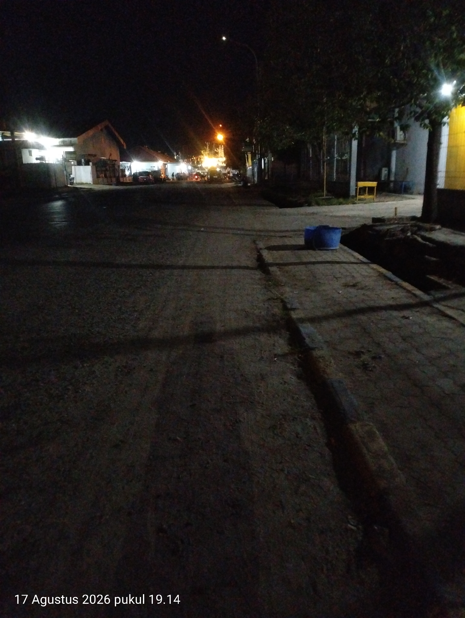 The image depicts a street scene at night in Kendari, Indonesia, with artificial lighting illuminating parts of the environment. On the left side of the road, several buildings with illuminated windows and doorways are visible, suggesting commercial or residential establishments. Some vehicles appear to be parked in front of these buildings. Trees line the right side of the street, casting shadows on the road. A sidewalk runs alongside the trees, and on it, two blue plastic buckets are placed. A yellow bench is also visible further down the sidewalk. The ground is paved, likely asphalt, and shows signs of wear and tear. The overall impression is of a quiet, dimly lit urban area after dark. The timestamp at the bottom of the image indicates the photograph was taken on August 17, 2026, at 7:14 PM.