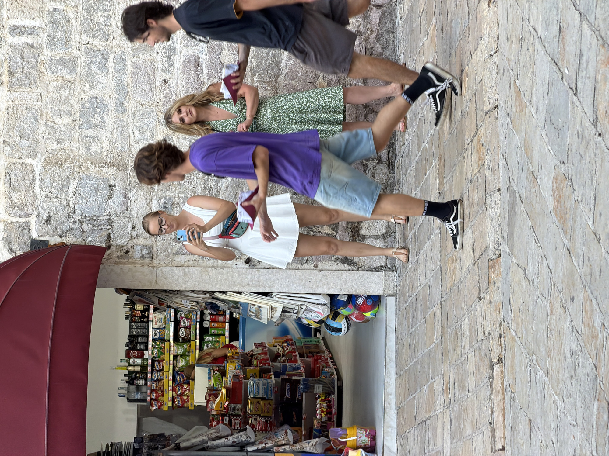 In Budva, Montenegro, on a cobblestone street, a group of four individuals is captured in a moment outside a small convenience store. The store features a red awning and displays shelves stocked with bottled drinks, snacks, and other merchandise.
On the left, a woman with blonde hair and glasses is holding a mobile phone, seemingly taking a picture. She is wearing a white dress and a fanny pack. To her right, a young man in a purple t-shirt and denim shorts walks away from the camera, holding a red object. Next to him, a woman with blonde hair in a green floral dress looks towards the camera, also holding a red object. On the far right, a man in a black t-shirt and dark shorts walks away from the camera, also holding a red object. The foreground is dominated by the textured cobblestone pavement. The background features a stone wall. The overall scene suggests a casual street encounter in a tourist area.