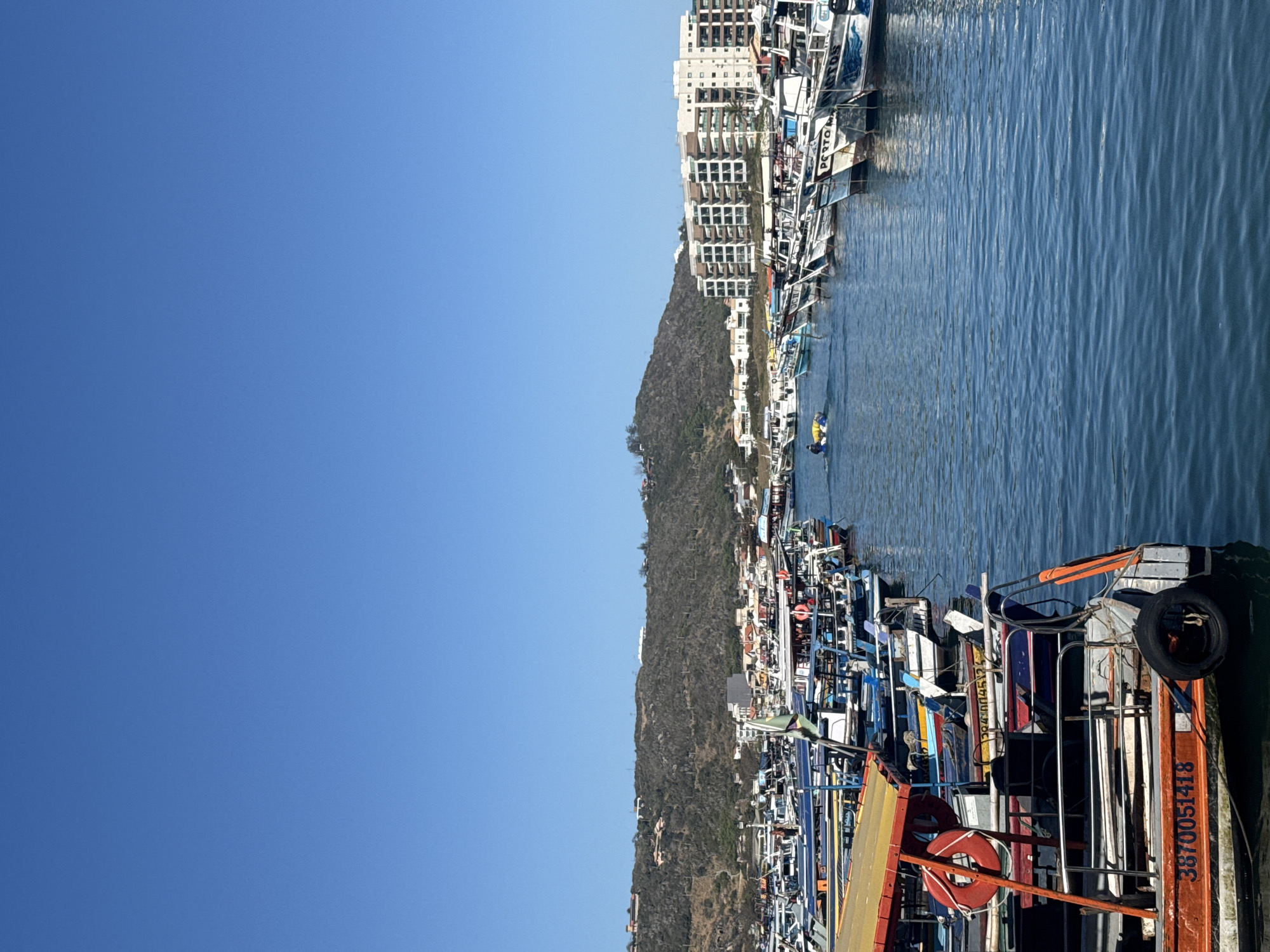 This is a photograph of a busy harbor in Arraial do Cabo, Brazil, under a clear blue sky. The foreground is dominated by a brightly colored boat, predominantly orange and yellow, with a life preserver and a number "3870051418" visible on its hull. Beyond this boat, a multitude of other vessels, primarily fishing boats and smaller passenger boats, are docked in neat rows. Many of these boats have colorful awnings and varying designs, indicating a diverse fleet. In the middle ground, a person is seen in a small yellow raft in the water, paddling.
The background features a densely populated shoreline with a mix of buildings, including what appears to be a large, modern apartment complex or hotel with a white facade and multiple balconies, situated on a hill. Behind the buildings, a rugged, tree-covered hill rises, also dotted with houses. The water of the harbor is a deep blue and appears calm, reflecting the clear sky and the surrounding boats and buildings. The overall impression is one of a vibrant coastal community and a working port.