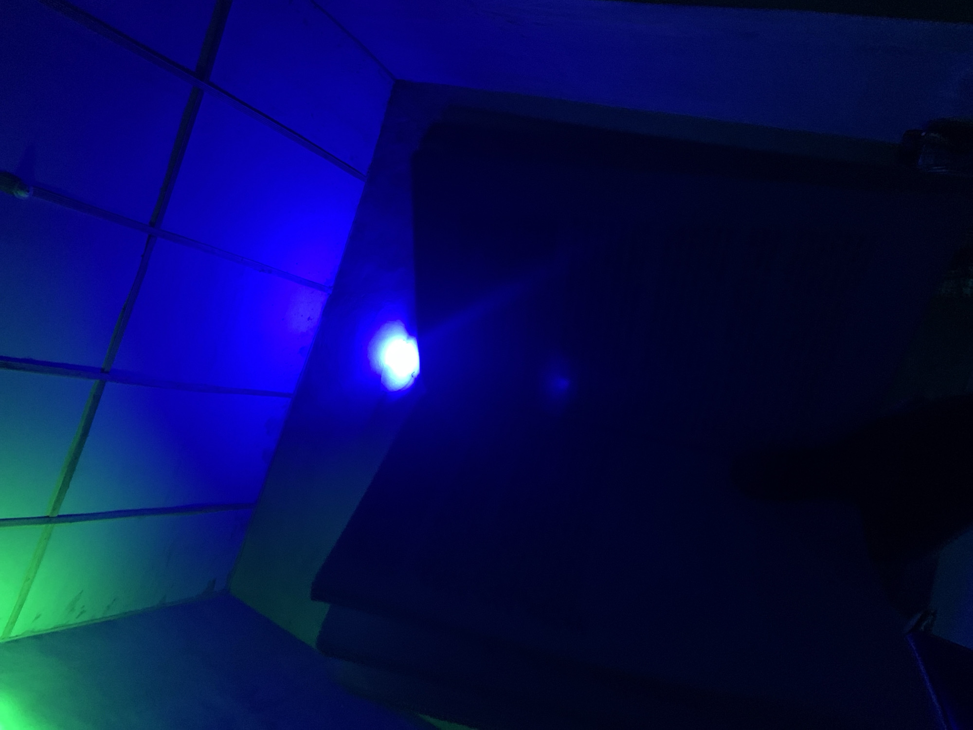 The image shows an interior scene illuminated by blue and green lights. The environment appears to be a room with a tiled ceiling that is lit with blue light. A large, dark surface, possibly a wall or screen, occupies the central and lower portion of the frame. A bright blue light source is visible near the top of this dark surface. Green light illuminates the upper left corner of the scene, suggesting another light source or the reflection of light from an adjacent area. The overall impression is one of low light conditions with artificial colored illumination. No people or specific identifiable objects are clearly visible. The location is unknown.