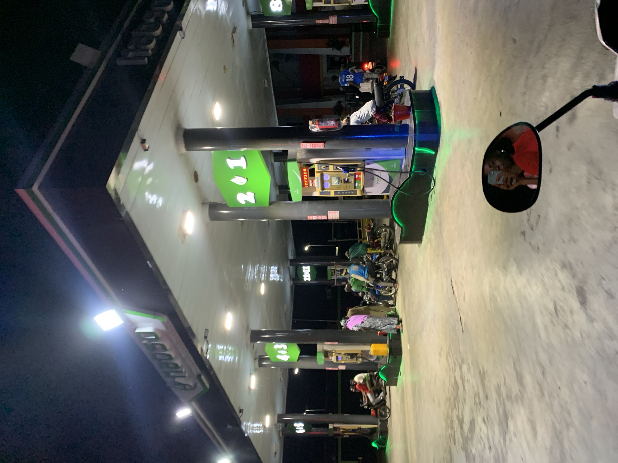A gas station in Lokoja, Nigeria, at night. Motorbikes line up at pumps numbered 2, 4, 6, and 8. The gas station's sign is visible above the pumps. A person is visible in a motorcycle mirror. Patrons are filling fuel.