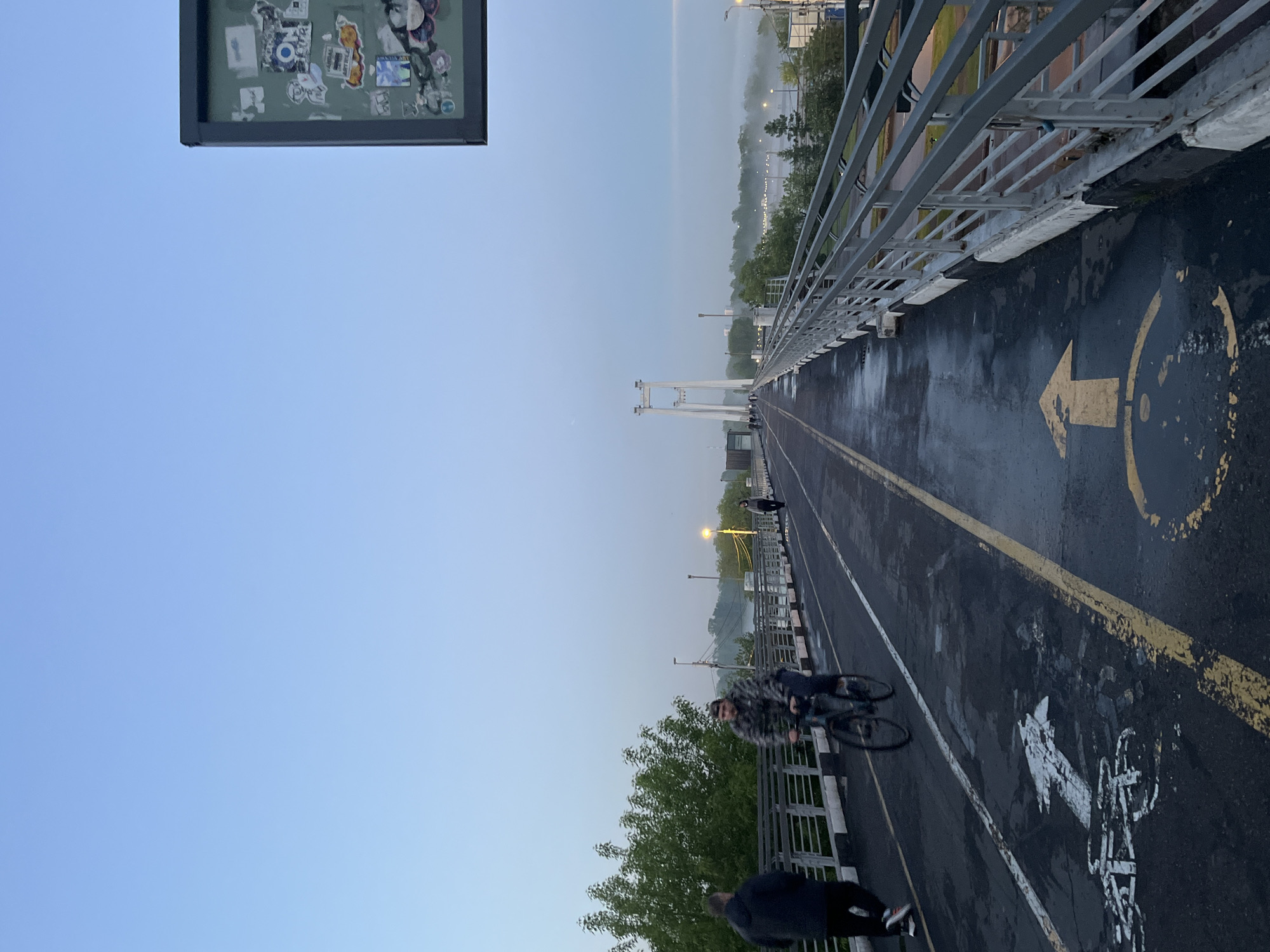 The image depicts a pedestrian and bicycle bridge in Krasnoyarsk, Russia, with a view looking lengthwise across the bridge. On the bridge surface, there are marked lanes with painted bicycle symbols and directional arrows. A cyclist is moving along one of the lanes. A pedestrian is walking on the edge of the bridge. Railings are visible along the sides of the bridge. In the background, there is a hazy skyline with trees and buildings. A sign with various stickers is at the left edge of the frame.