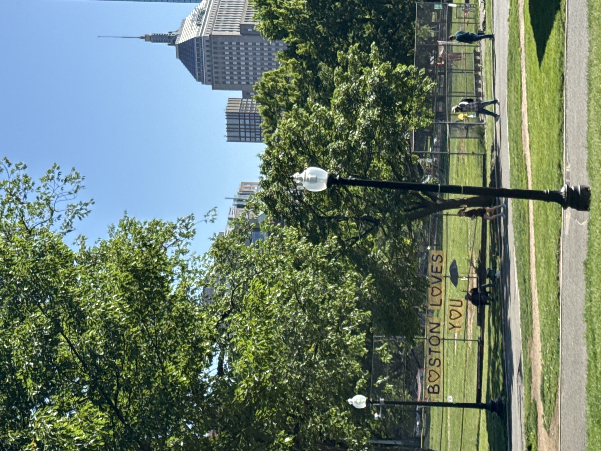 The image captures a vibrant urban park scene in Boston on a clear, sunny day. Lush green trees, with their leaves illuminated by bright sunshine, frame the foreground and midground. In the background, the distinctive skyline of Boston is visible, featuring several tall buildings under a brilliant blue sky, including one with a prominent spire, characteristic of the Custom House Tower.
A black, ornate street lamp stands prominently on the right side of the frame, with another similar lamp visible further left. A paved path runs alongside a well-kept lawn, with a chain-link fence acting as a divider within the park. Attached to this fence is a white sign with red lettering that prominently displays the text "BOSTON ❤️ YOU," clearly identifying the location and conveying a welcoming sentiment.
Several individuals are enjoying the park. One person, wearing a plaid shirt, walks along a path, while another is seen further in the distance. Other figures are scattered across the grassy areas, some walking, others potentially seated on benches behind the fence. The bright conditions, clear sky, and strong shadows indicate it is daytime, likely late morning or afternoon, with excellent weather for outdoor activities. The overall atmosphere is peaceful and inviting, reflecting the positive message on the sign.