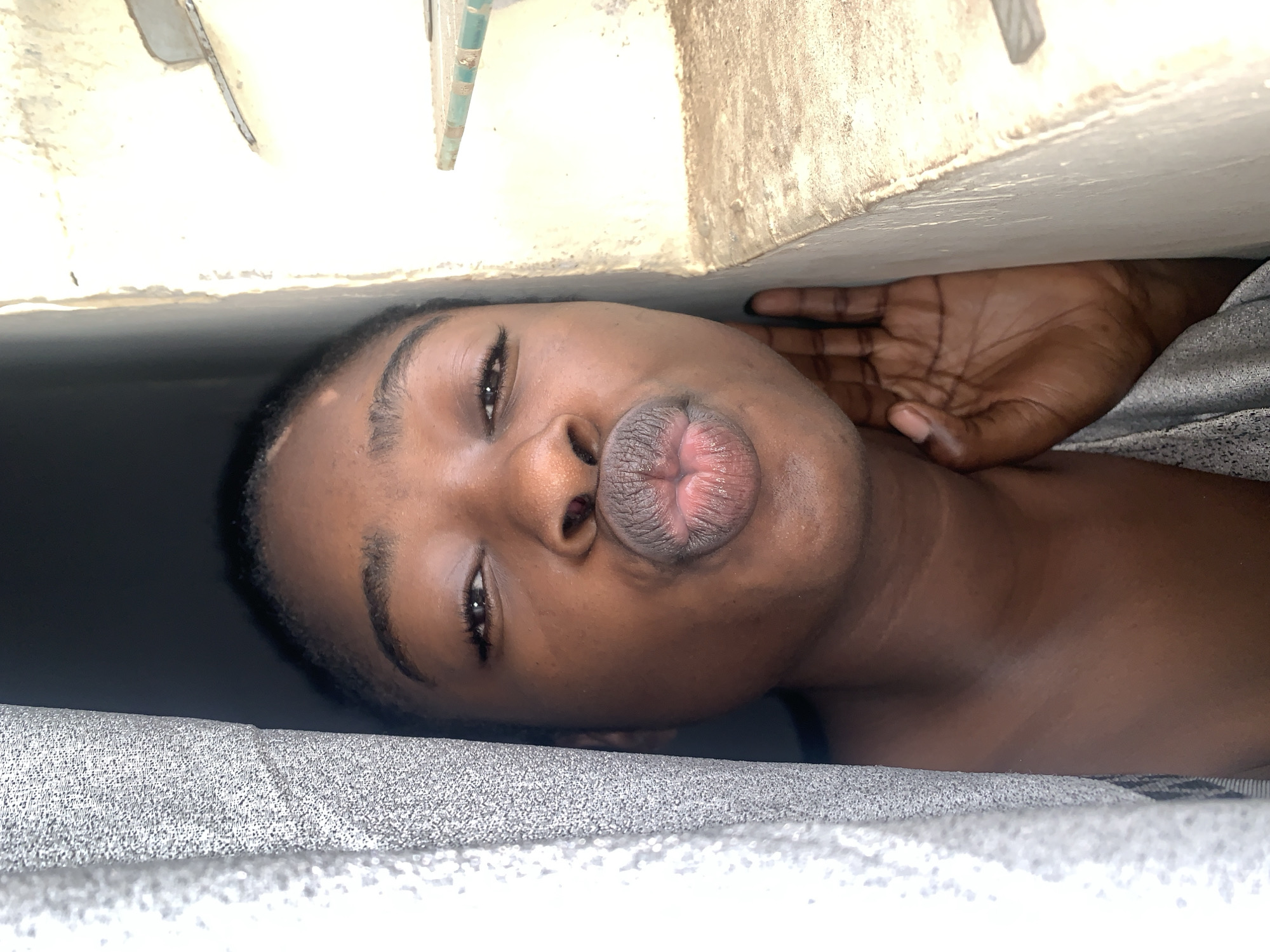 A young Black woman is pictured in a close-up shot, her face partially obscured by a grey curtain on the left and a beige wall on the right. She is looking directly at the camera with her lips puckered for a kiss. Her right hand is visible on the right side of the image, pressed against the wall. The background on the right shows a weathered wall with a ledge and some green trim. The lighting suggests an outdoor or well-lit indoor environment. The image appears to have been taken in Rafin Par, Nigeria.