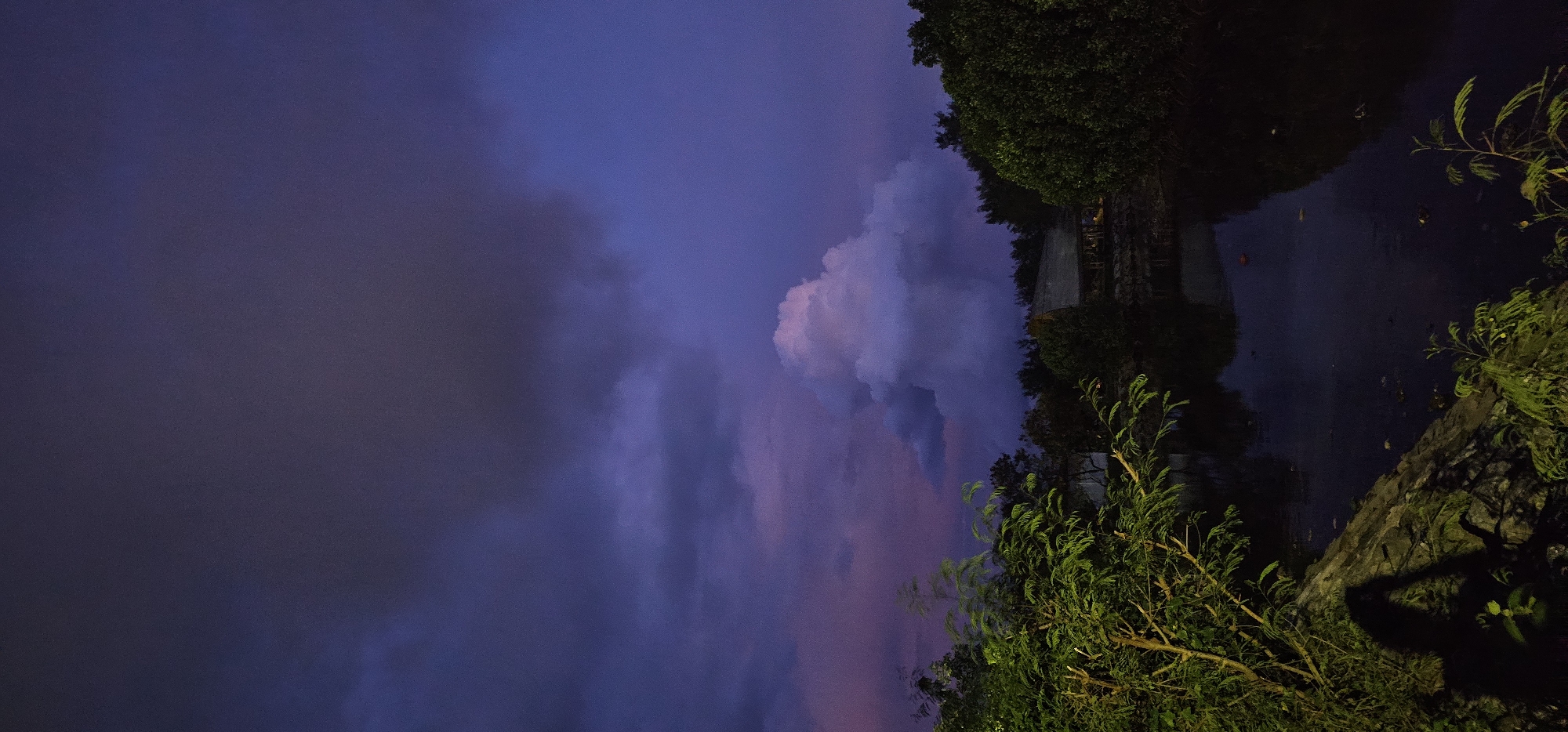 The image captures a serene, twilight landscape, likely at dusk, with a rich palette of deep blues, purples, and blacks dominating the scene. The sky, taking up the upper left portion of the frame (when viewed upright), is filled with large, dramatic clouds. Some clouds exhibit soft pink and white highlights, suggesting the lingering light of a setting sun, while others are darker grey and purple, blending into the darkening sky.
In the mid-ground and foreground, lush green foliage and trees line what appears to be a body of water, possibly a canal or small river, which occupies the lower right section of the image. The water's surface is dark and reflective, mirroring the deep blue and black tones of the sky and the silhouettes of the surrounding greenery and structures. On the bank to the right, some rocks or a low wall are visible, partly covered by vegetation.
Faintly visible through the trees on the opposite bank (in the upper right when upright) are the dark outlines of buildings or structures, adding an element of urban or semi-urban environment to the natural landscape. The overall lighting is low, creating a mysterious and calm atmosphere. The dense, dark green of the foliage, consistent with tropical environments, subtly hints at the Denpasar, Indonesia location context. No discernible activity is taking place, and no text is visible in the image.