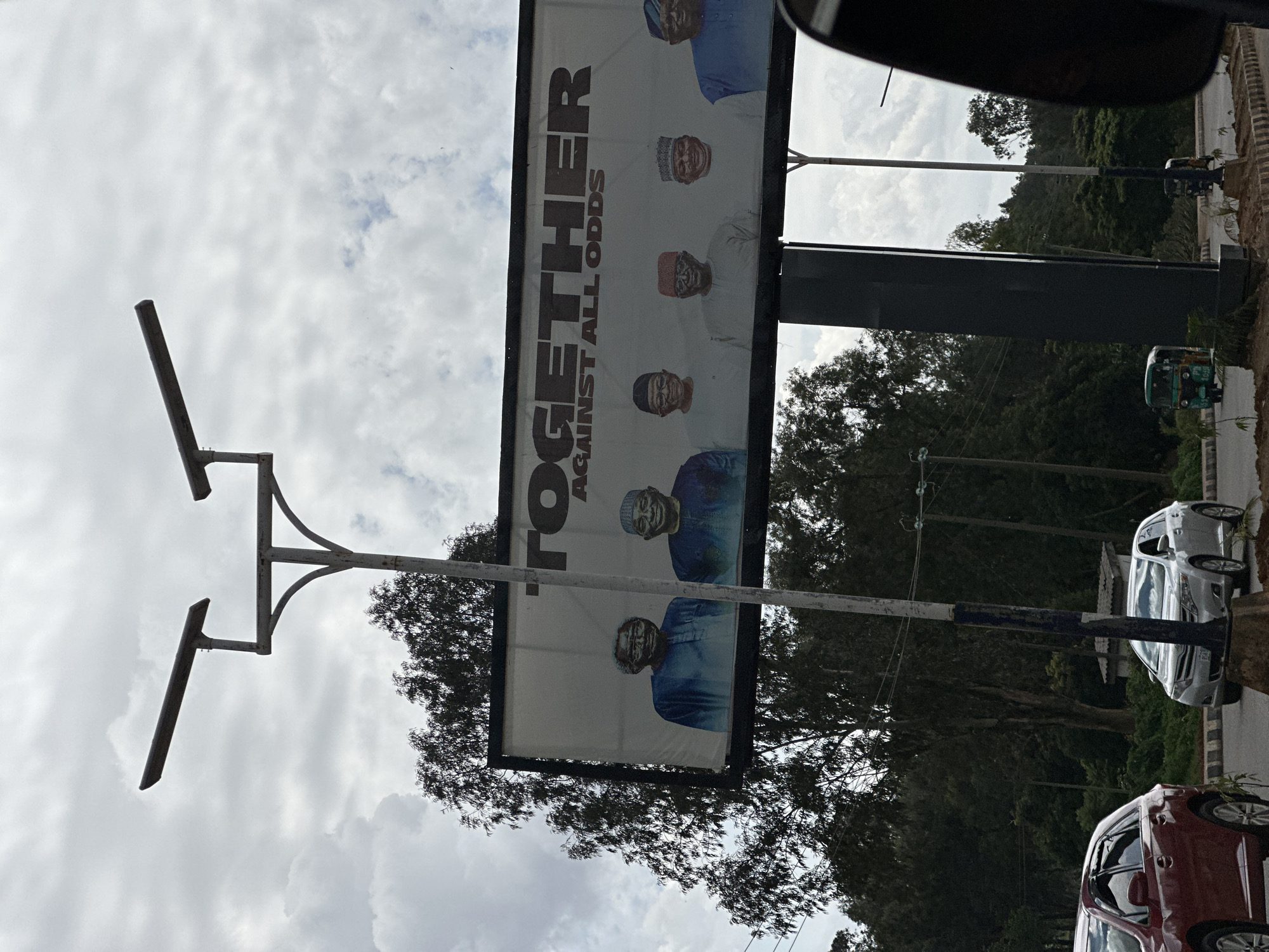 A billboard displaying the word "TOGETHER" in large, bold, black letters is positioned vertically in the center of the image. Below this, in smaller text, it reads "AGAINST ALL ODDS". The billboard features portraits of five men, all wearing traditional Nigerian attire and caps. The men appear to be smiling or in a neutral expression.
The billboard is mounted on a large metal pole structure, which is also oriented vertically. In the background, there are trees and a cloudy sky. The image is taken from a low angle, looking up at the billboard.
To the right of the billboard, there is a road with several vehicles. A small, green three-wheeled vehicle (possibly an auto-rickshaw) is visible further down the road. Two cars, one silver and one red, are parked on the side of the road. The overall scene suggests an outdoor setting, likely in a public area. The lighting indicates it is daytime, and the cloudy sky suggests overcast weather. The presence of the billboard with political or campaign-like imagery, combined with the traditional attire of the men, hints at a political or social event taking place.