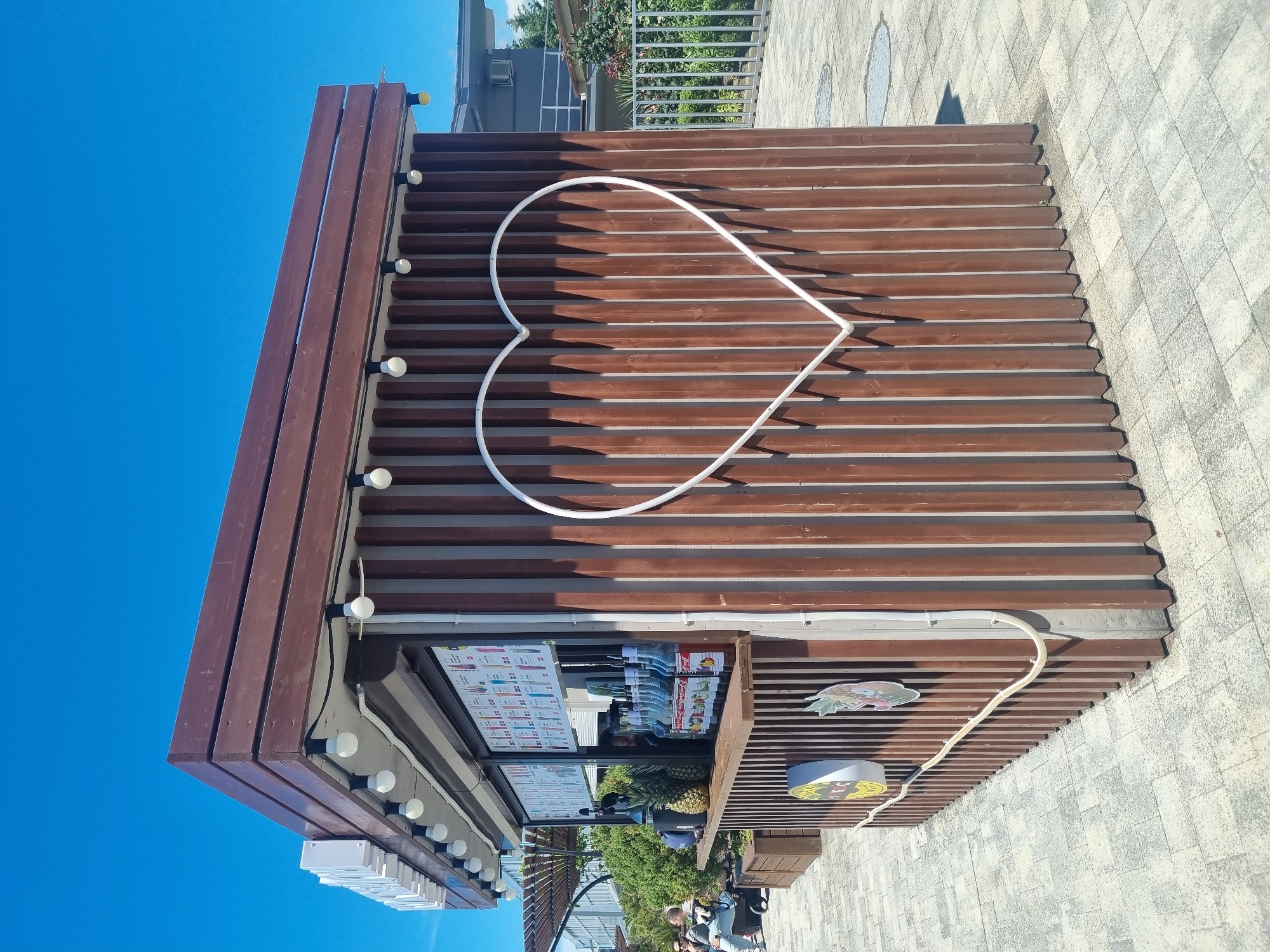 The image captures a street food kiosk in Sochi, Russia, under a clear blue sky. The kiosk has a brown wooden facade with a large white heart-shaped neon light on its side. It sells refreshments as indicated by the menu boards displayed and the arrangement of goods. Below, there are people gathered on the street, including someone who seems to be seated and several standing near the kiosk. The surrounding area appears to be a pedestrian zone with tiled paving.