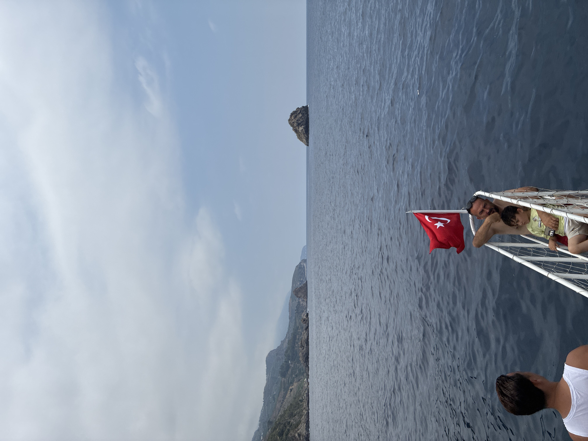 The image depicts a view from a boat at sea, with the coastline of Gazipaşa, Turkey, visible in the distance. The water is a deep blue, with gentle waves. A rugged coastline with green vegetation and rocky outcrops stretches across the left side of the frame. In the middle distance, a small, rocky island rises from the sea. The sky is overcast with scattered clouds.
In the foreground, on the deck of the boat, are two people. A man, shirtless and wearing sunglasses on his head, is seated and holding a young child. The child is wearing a light-colored t-shirt and shorts. Beside them, a Turkish flag with a white star and crescent on a red background flutters from a pole. To the left of this group, a man from the shoulders up is visible, wearing a white tank top, looking out towards the sea. The boat has a white railing.