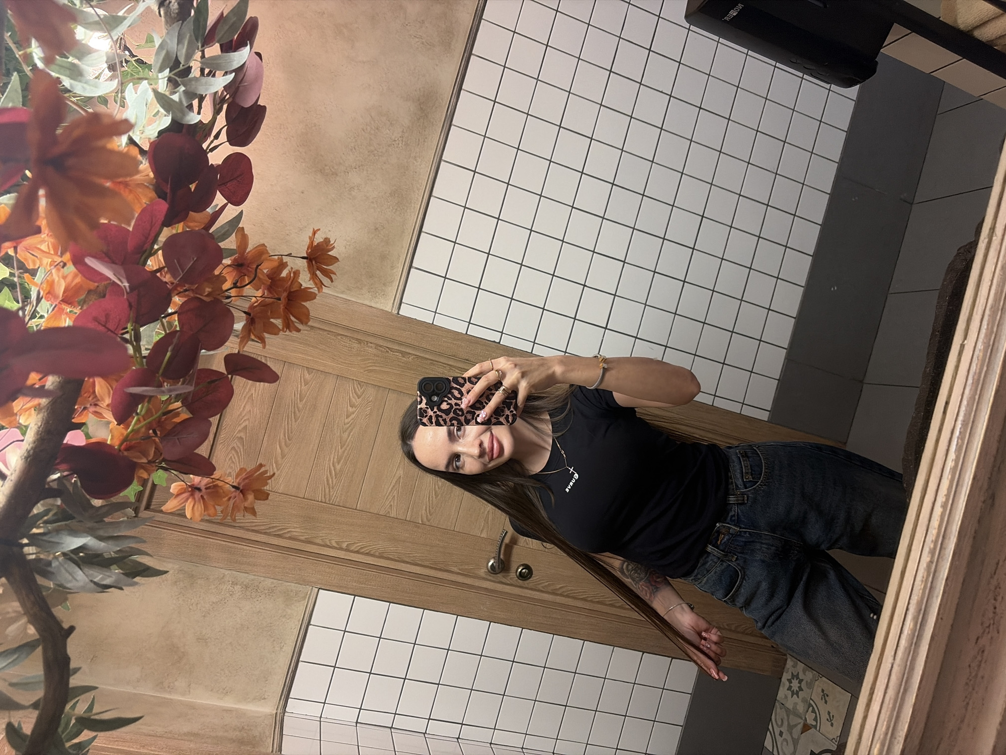 A young woman is captured taking a mirror selfie indoors, likely in a public restroom or washroom, in Izhevsk, Russia. She holds a smartphone with a leopard print case up to capture her reflection, smiling faintly at the camera.
The woman has long, straight dark hair and is dressed in a black short-sleeved t-shirt with partially visible white text that appears to read "BVRU+". She is also wearing blue jeans. Rings adorn her fingers, a bracelet is on her wrist, and a tattoo is partly visible on her left forearm.
The background features a mix of white square tiles with dark grout lines on the lower walls and a textured light brown/beige wall above. A wooden door or paneling is visible behind the woman. To the left, a prominent display of decorative artificial plants with autumn-colored leaves in shades of red, orange, and green-grey adds warmth to the setting. In the upper right corner, a black wall-mounted device, possibly a hand dryer or soap dispenser, is visible, with the text "MOIBEH." printed on its side. The lighting suggests a well-lit indoor environment.