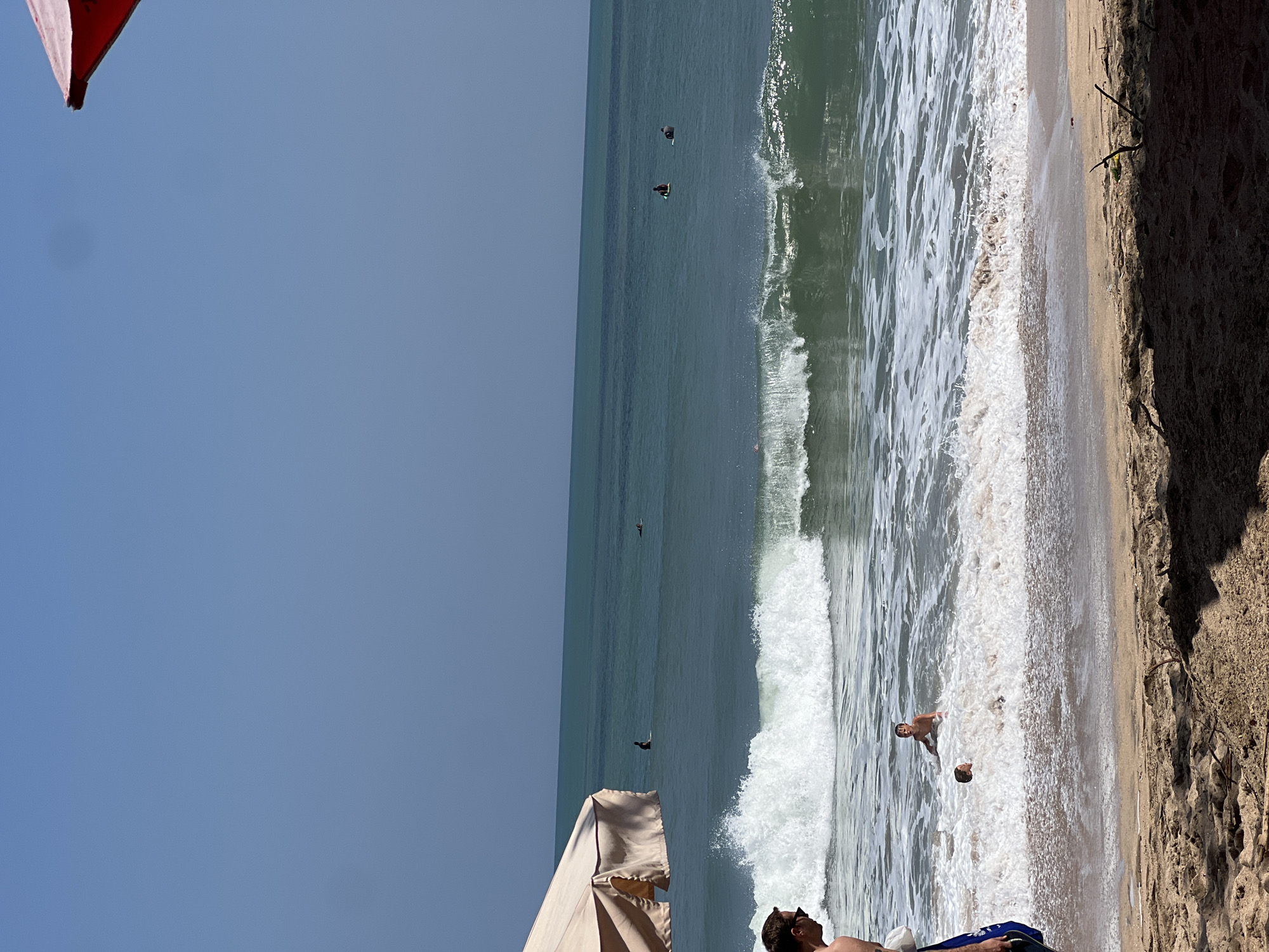 In Kuta, Indonesia, on a sunny day at the beach, the ocean is a deep teal, with a clear horizon under a pale blue sky. Gentle waves break on the shore, creating white foam that washes over the sand. Several surfers are visible in the distance, seated on their boards. Closer to the beach, two young boys are playing in the surf, splashing and laughing. In the foreground, a person is standing on the sand, facing away from the camera, with a large umbrella providing shade. Another person is partially visible to the left, wearing dark sunglasses. The beach sand is a light brown, with some debris scattered on it.