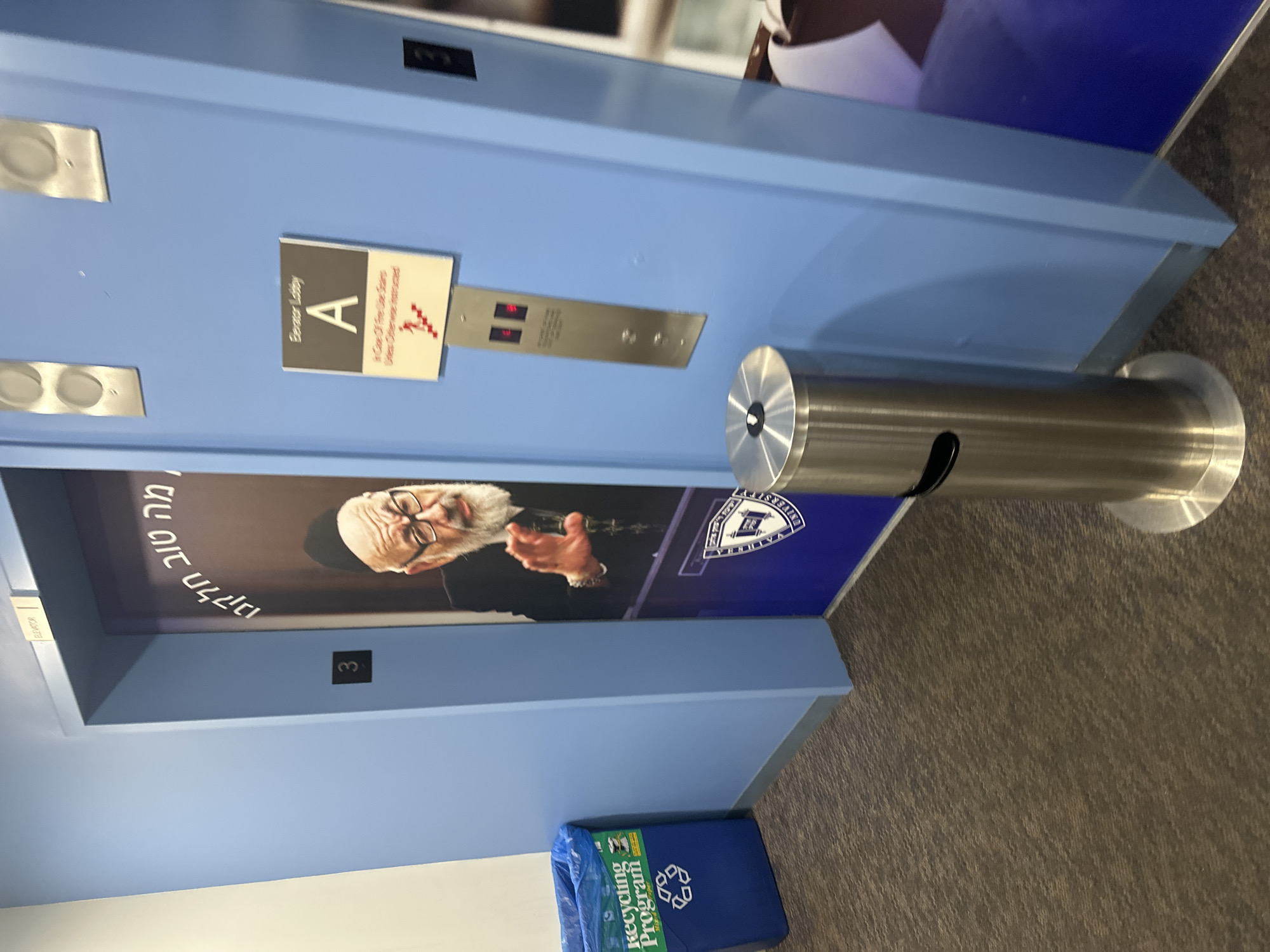 Here is a description of the image:
The image shows a building interior, specifically an elevator area with elevator signs. A sign indicates it's "Elevator Lobby A" and includes instructions to "In Case Of Fire Use Stairs Unless Otherwise Instructed". Below the elevator button panel, which indicates floors "3" and "5", there is a poster with a photograph of a man wearing a black skullcap and glasses. The poster includes writing in Hebrew and the logo for Yeshiva University.
A stainless steel trash receptacle is placed next to the elevator bank. On the floor, there is a blue recycling bag labeled "Recycling Program." The color scheme of the elevator doors is light blue. The floor is carpeted in what appears to be a dark brown or gray carpet. The photo was likely taken indoors in a commercial or institutional setting, possibly in a building in the city of New York, United States of America, where Yeshiva University has its main campus.