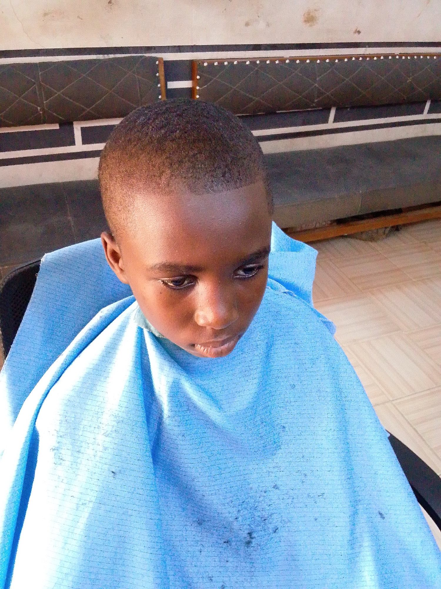 A young child, likely a boy with dark skin, is seated indoors, possibly in a barbershop or salon, wearing a light blue barber's cape. The child's hair is very short and neatly trimmed, with a sharp, clean hairline around the forehead, suggesting a recent haircut. The child is looking down and slightly to their right, with a neutral to slightly pensive expression.
The light blue cape covers the child's shoulders and torso, showing some dark specks, possibly hair clippings, particularly on the lower part. The child is seated in a dark-colored chair, of which only parts of the frame are visible.
In the background, a dark, quilted sofa or bench with decorative studs along its top edge is visible. Behind this seating, a wall features horizontal black and white stripes and a lighter-colored section above. The floor is covered with light-colored square tiles. The lighting suggests it is daytime, casting a bright illumination on the scene. There is no visible text.