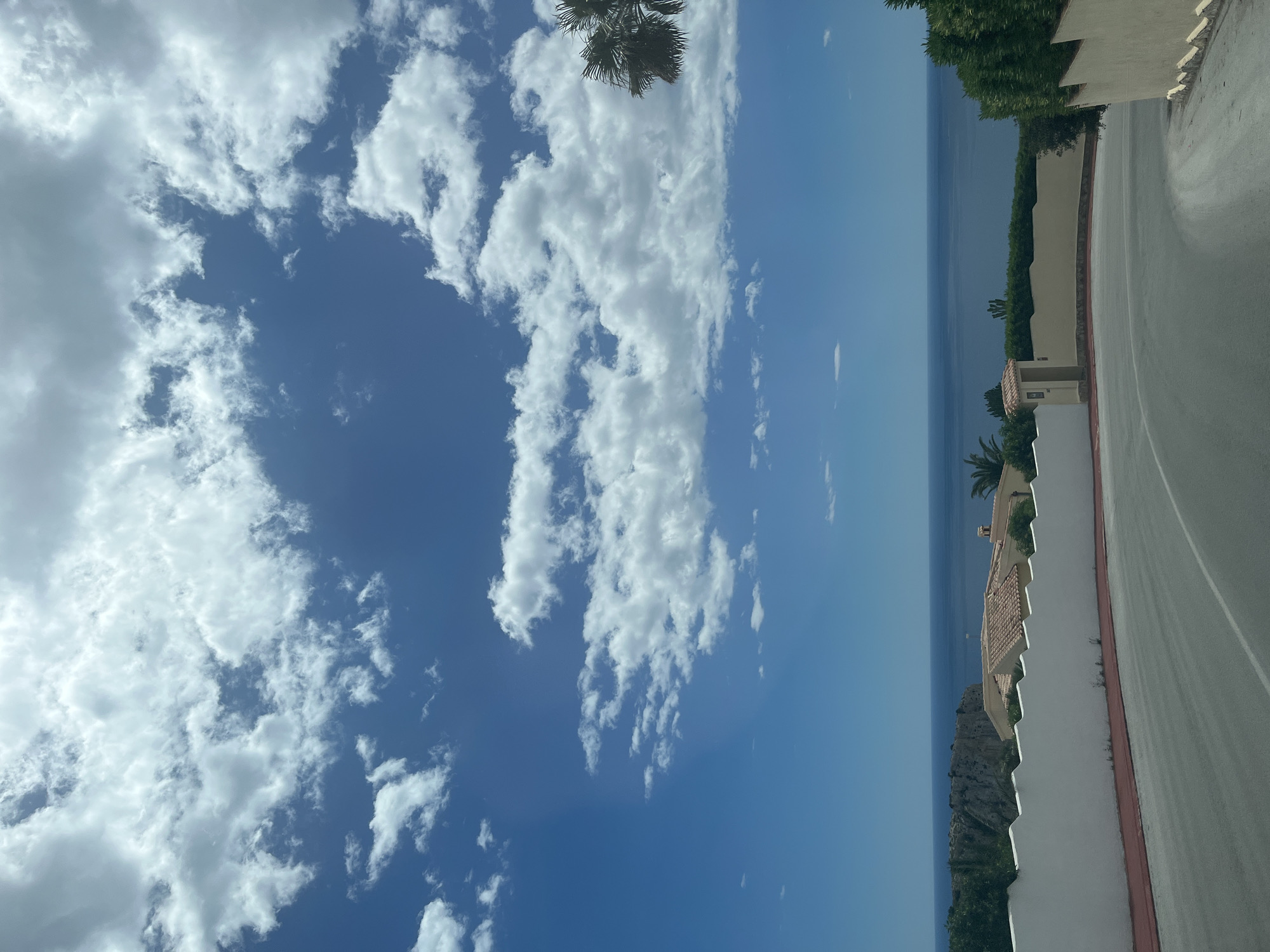 The image shows a view of Urbanització El Cim Del Sol, Spain, on a partly cloudy day. The sky is mostly blue with numerous white cumulus clouds covering a large portion of it. In the distance, there is a view of the Mediterranean Sea, appearing as a horizontal blue band. A road curves into the right side of the frame, lined by a white wall with a red trim at the bottom. Vegetation and a few houses are visible on the other side of the wall. A single palm tree frond is visible near the top of the image.