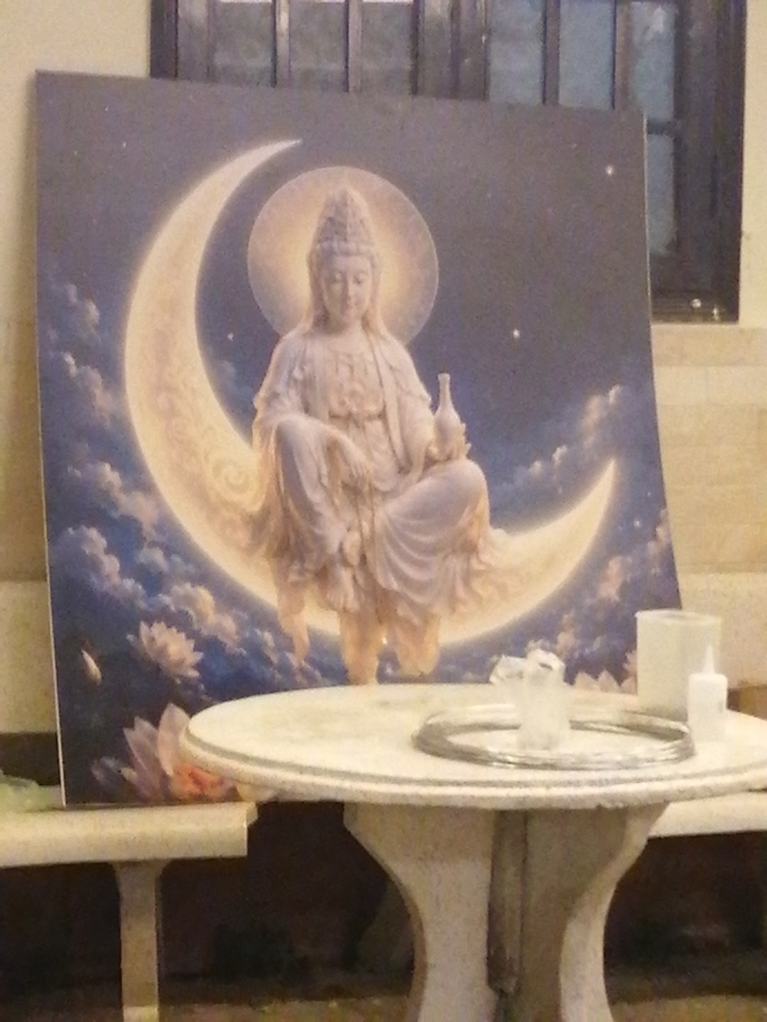 The image shows a painting of a deity, possibly Guanyin or a similar figure, seated on a crescent moon against a night sky background. The painting rests on a bench behind a round, marble-topped table. On the table are a silver ring, small glass objects, and a white candle and a small white bottle. The scene appears to be indoors, with a barred window visible behind the painting. Based on the presence of Buddhist imagery, it is likely this image was captured in Da Lat, Vietnam, which is known for its many temples and shrines.