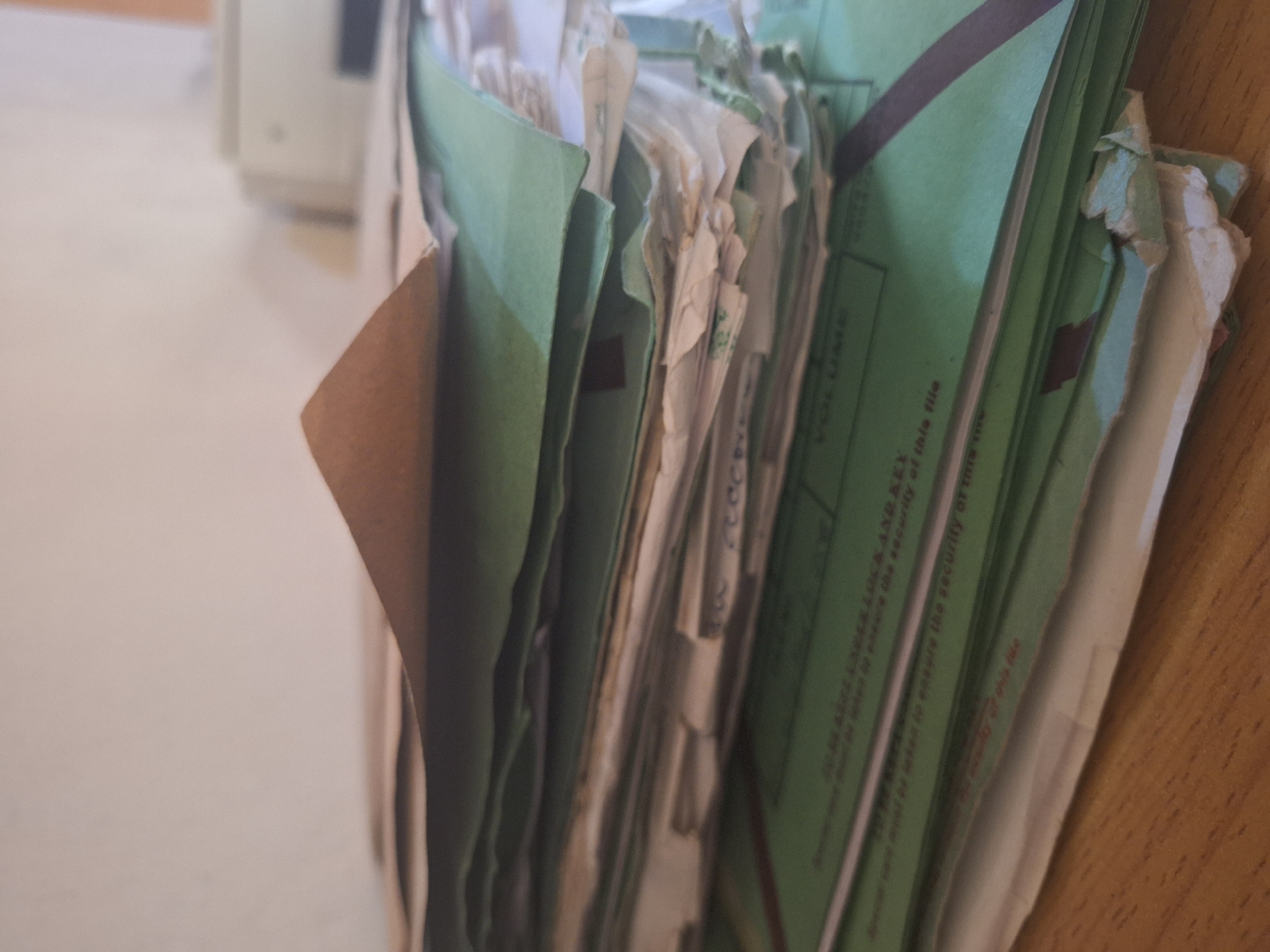 A stack of papers and folders is piled on a wooden desk. The papers are mostly white and cream-colored, with some green folders mixed in. Some of the papers are yellowed and appear to be old. The stack is leaning to the left, with a brown paper folder peeking out from the top. The background is a light-colored wall. The image is taken in Jalingo, Nigeria.