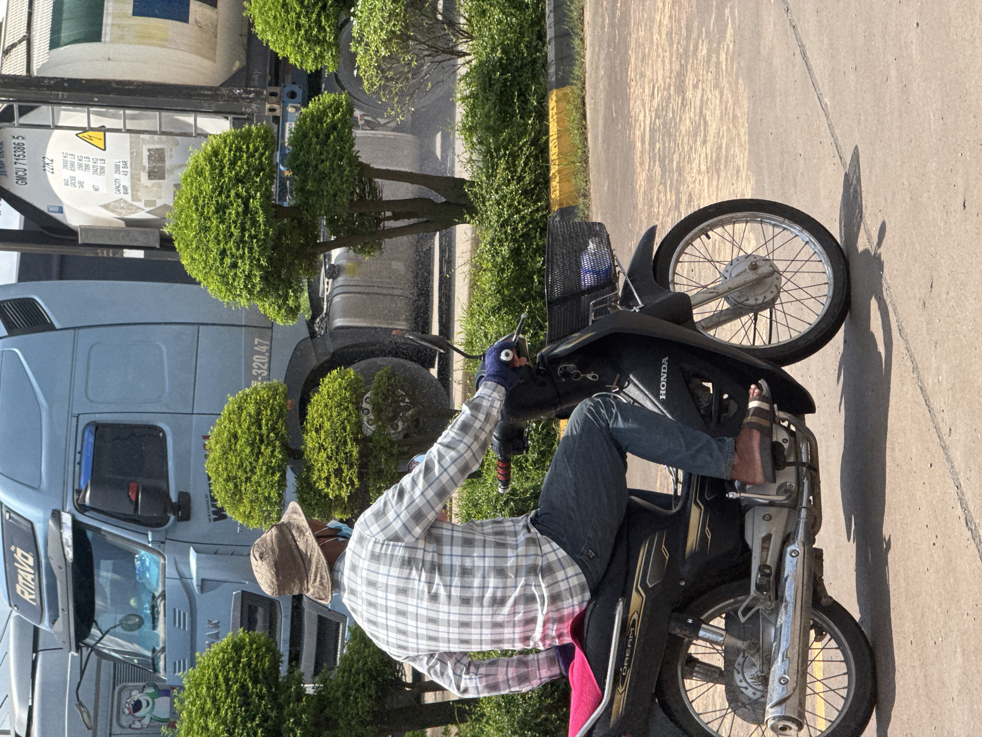 A man wearing a plaid shirt, blue jeans, a bucket hat, and sandals rides a black motorcycle in Bavet Quarter, Cambodia. He has a face mask on. The motorcycle has a pink seat cover and a black basket attached to the handlebars, which contains a white item. Behind the motorcycle is a large blue truck and a white tanker trailer. The ground is paved concrete, with a yellow and black striped curb visible on the right. There are green bushes and trees in the background. The scene appears to be outdoors on a sunny day.