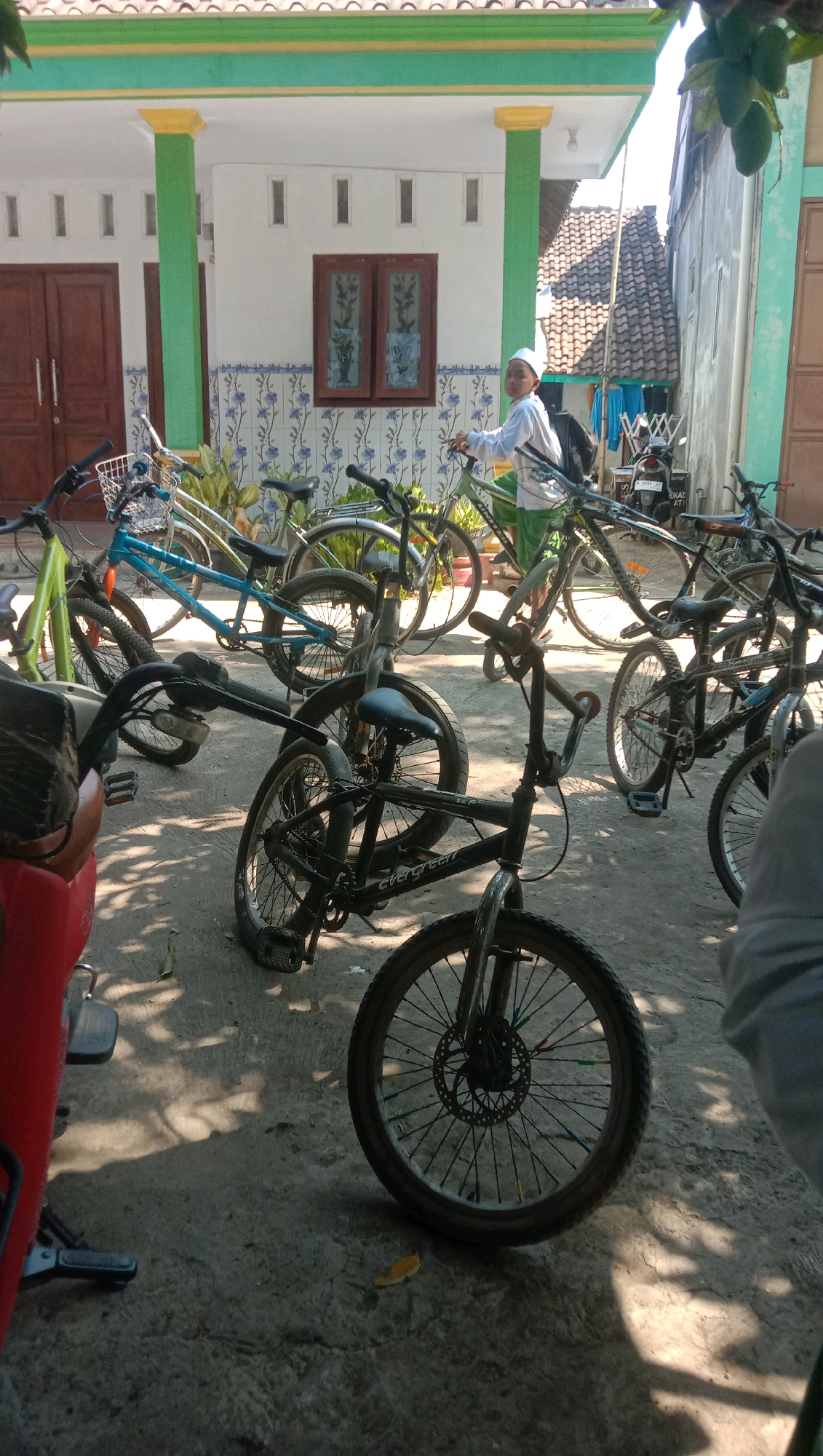 In Ngempit, Indonesia, on a sunny day, a young boy wearing a white cap and traditional clothing stands next to a green bicycle. He is positioned in front of a building with white walls, green trim, and yellow accents on the columns. The building has a dark brown double door and a window with decorative glass. Several other bicycles are parked nearby, including a blue one, a black BMX-style bike, and a lime green bicycle. The ground is paved with concrete, and shadows from trees are cast across it. In the background, a motorcycle is parked near a tiled roof.