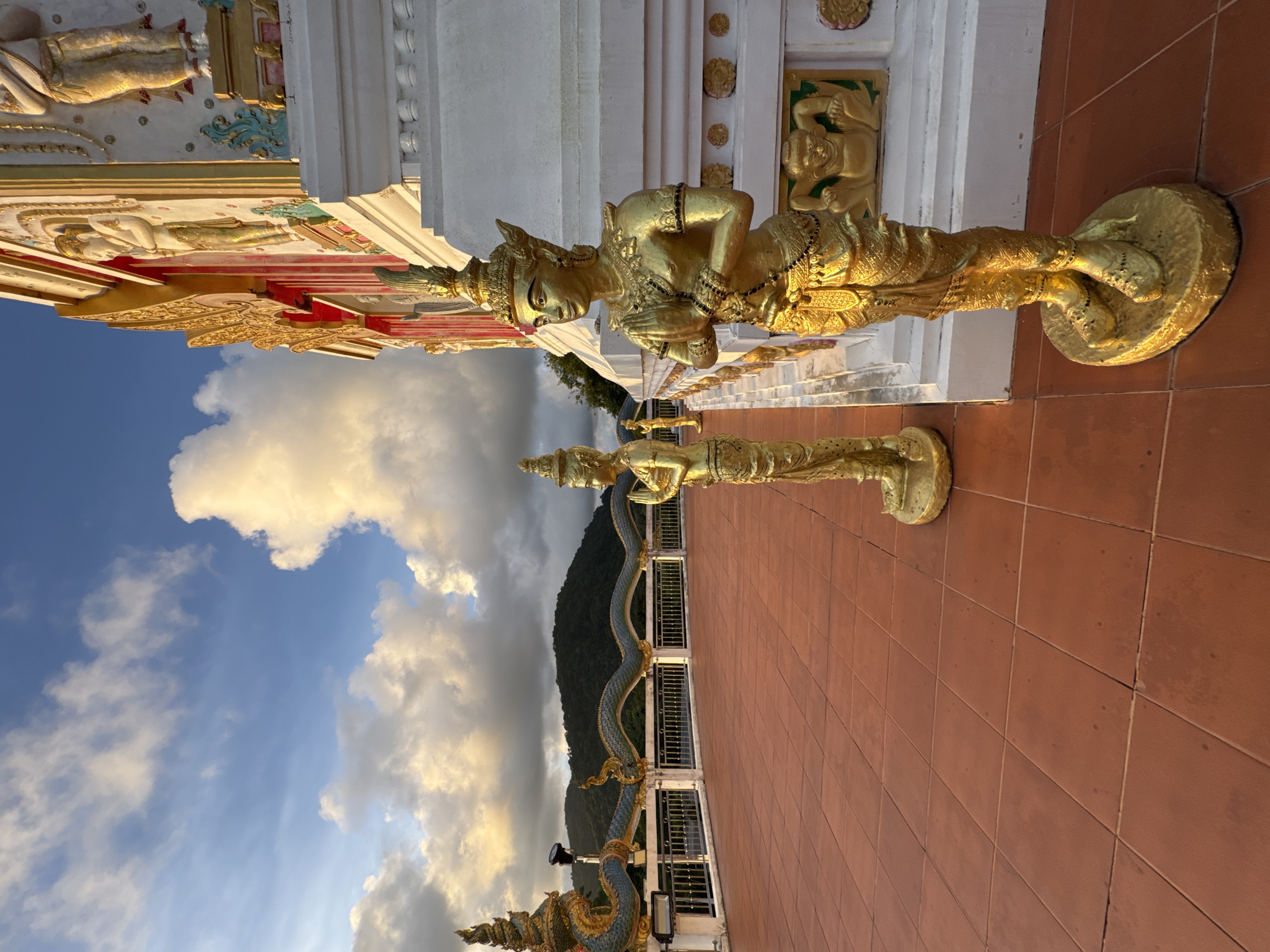 This is a photograph taken at a temple in Patong, Thailand, during what appears to be late afternoon or early evening. The foreground is dominated by a wide, reddish-orange tiled plaza. To the right, a golden statue of a person with hands pressed together in a gesture of prayer stands prominently. This statue is adorned with intricate jewelry and a headdress. Further into the plaza, another similar golden statue is visible, positioned behind and to the left of the first.
In the background, the architecture of the temple is a focal point. It features white walls with ornate gold and red decorative elements. A section of a decorative railing, possibly topped with a golden serpent motif, runs along the edge of the plaza. Beyond the temple, a lush green mountain range is visible under a sky filled with dramatic clouds. The clouds are illuminated by the setting sun, creating a warm, golden glow. The overall impression is one of serene beauty and cultural richness.