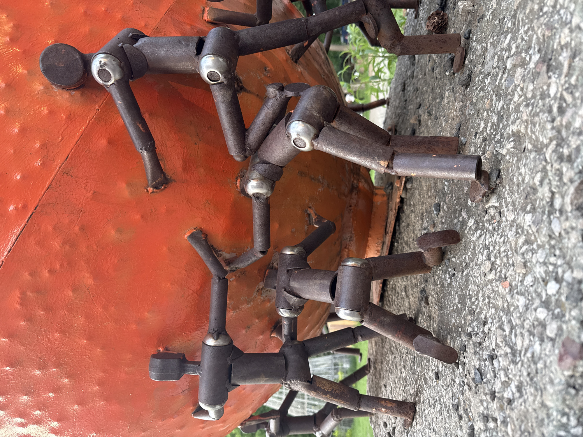 The image shows a close-up of a metal sculpture, likely located in Izhevsk, Russia, given the request's context. The sculpture consists of an orange, rounded object to which numerous metal rods and cylinders are attached. These metal components appear to be arranged in a repetitive pattern, possibly representing stylized human figures or abstract forms. The metal has a weathered, rusty appearance. The sculpture is situated on a concrete surface with a rough, textured finish. Background elements include hints of greenery, suggesting an outdoor setting, and a portion of a fence.