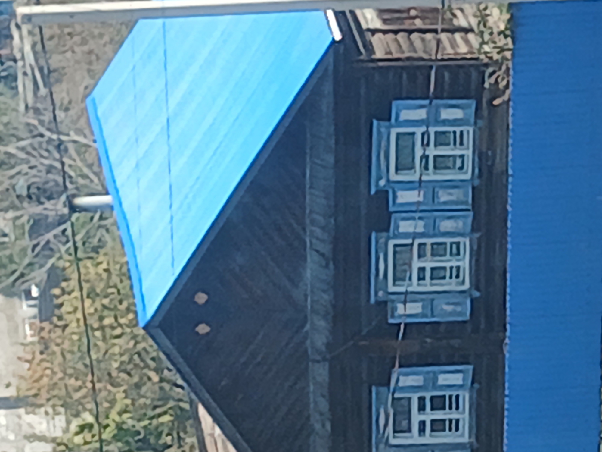 A close-up, eye-level shot shows the upper facade of a dark wooden house with a bright blue metal roof in Makinsk, Kazakhstan. The house has a gable roof with two circular, light-colored decorations on the upper part of the facade. Below the roofline, there are three windows with light blue shutters and white trim. The windows are divided into multiple panes. A portion of a blue fence is visible in the foreground. The background consists of blurred trees and foliage.