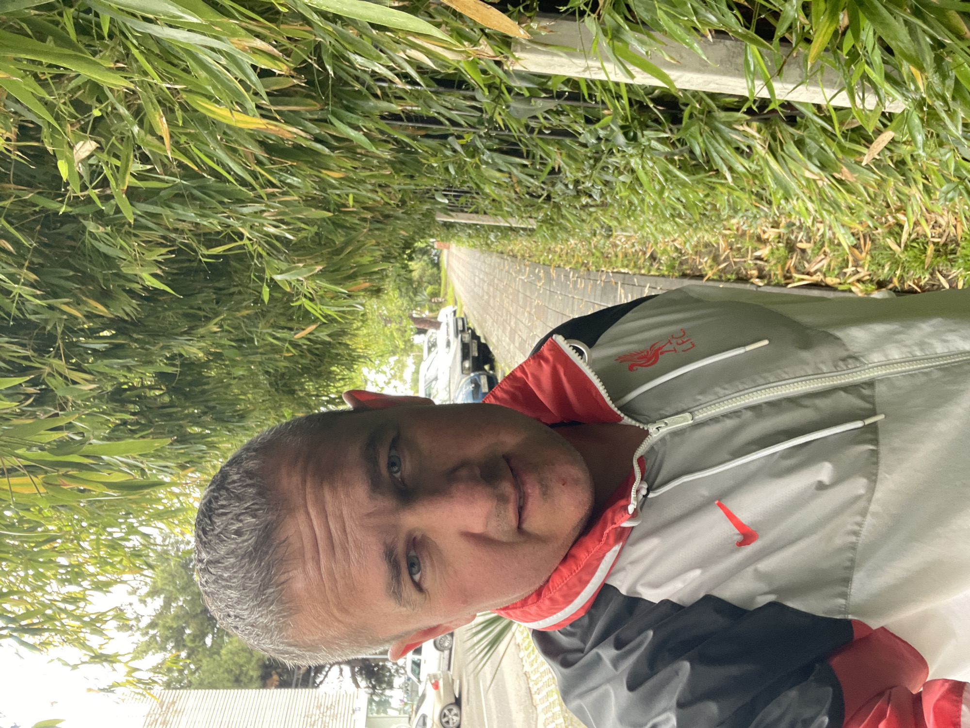 A man is standing on a brick walkway lined with bamboo plants on both sides. Beyond the plants are cars parked along a road and buildings. The man is wearing a jacket with gray, white, black, and red colors. A red Nike logo is visible on the jacket. A Liverpool FC logo is on the top left side of the jacket. Based on the greenery, architecture and vehicles, this could be an urban area in Sochi, Russia.