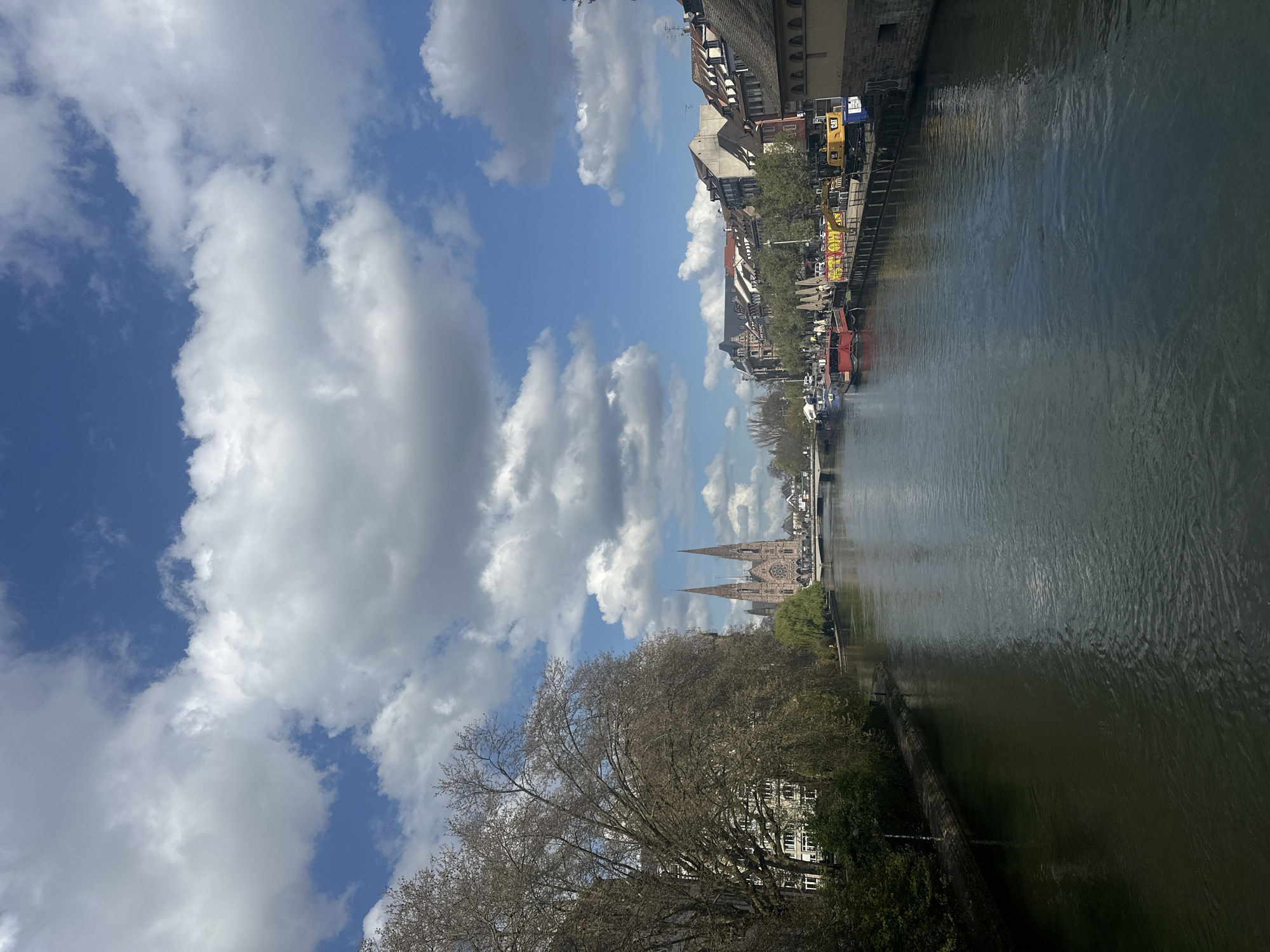 The image captures a view in Strasbourg, France, looking along a canal. The sky is bright blue, filled with large cumulus clouds. Along the right edge of the canal, there are buildings typical of Strasbourg architecture, some with timber framing, as well as trees and various boats docked along the waterway. A crane or some heavy equipment is also visible on the bank. In the distance, the spires of a cathedral rise above the cityscape. In the foreground, on the left bank of the canal, there's a large tree with bare branches and some buildings nestled behind it. The water of the canal is a dark, reflective green.