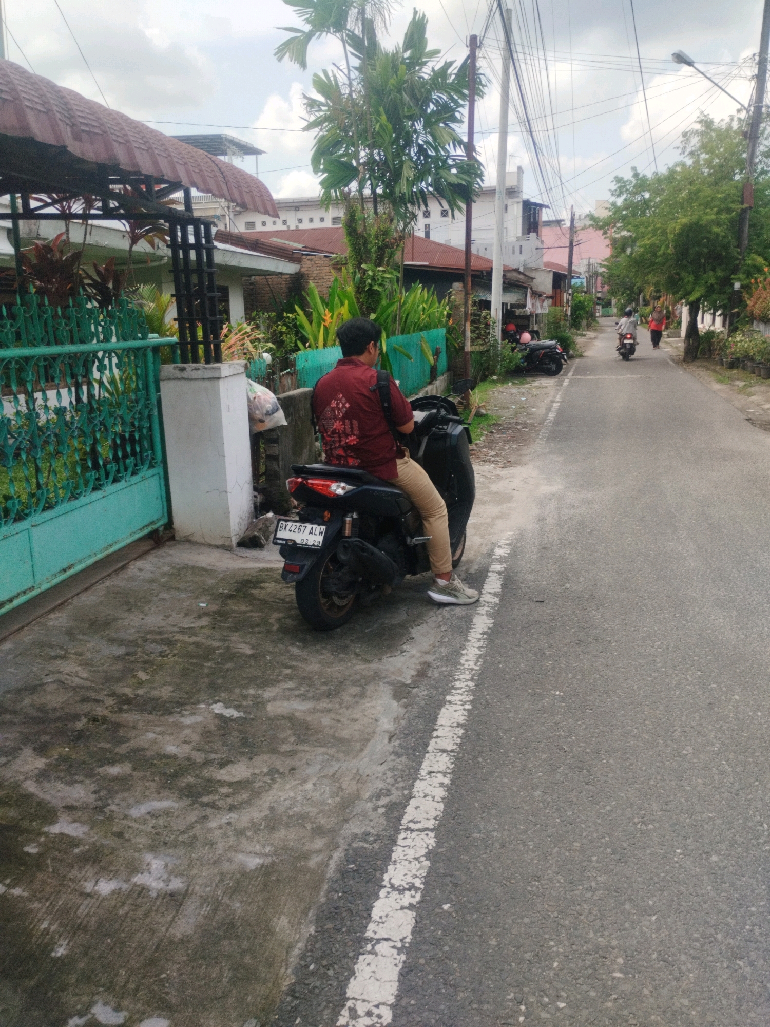 In the city of Medan, Indonesia, a person in a maroon shirt and tan pants sits on a black motorcycle parked on the side of a paved road. The motorcycle has a license plate that reads "BK 4267 ALW 03 23". The person is facing away from the camera. To the left of the motorcycle is a turquoise metal gate and a concrete pillar. Beyond the gate, a building with a brown, corrugated metal roof is visible. The road is lined with trees and other buildings. In the distance, two other motorcycles are seen on the road, one with a rider and another with a passenger. Overhead, power lines crisscross the sky. The ground on the left side of the road is a mix of concrete and asphalt, with some patches of vegetation. The weather appears to be overcast.