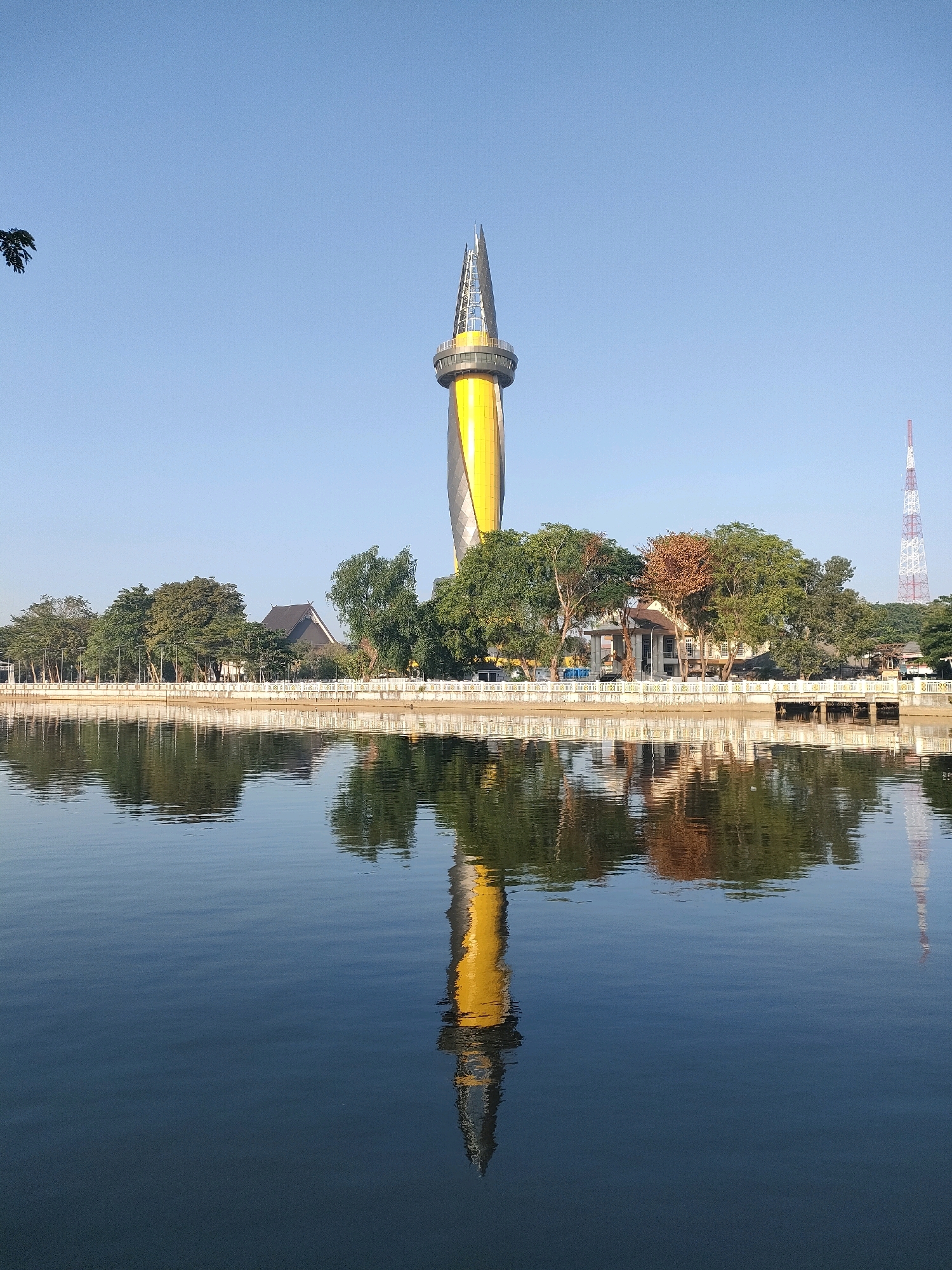 The image depicts a scenic view of the Bekantan Tower in Banjarmasin, Indonesia, reflected in a calm body of water. The tower, characterized by its striking yellow and black design, stands tall against a clear blue sky. It has a distinctive pointed top with visible structural elements. In the background, a line of trees and some buildings create a visual border between the water and the sky. One building appears to be a traditional-style house with a dark, pointed roof. A red and white lattice tower is also visible in the distance, to the right of the Bekantan Tower. The water's surface is smooth, creating a near-perfect mirror image of the tower, trees, and other structures. The lighting suggests it is daytime, likely morning or late afternoon, given the clear sky and the quality of the reflections. There are no people visible in the frame.