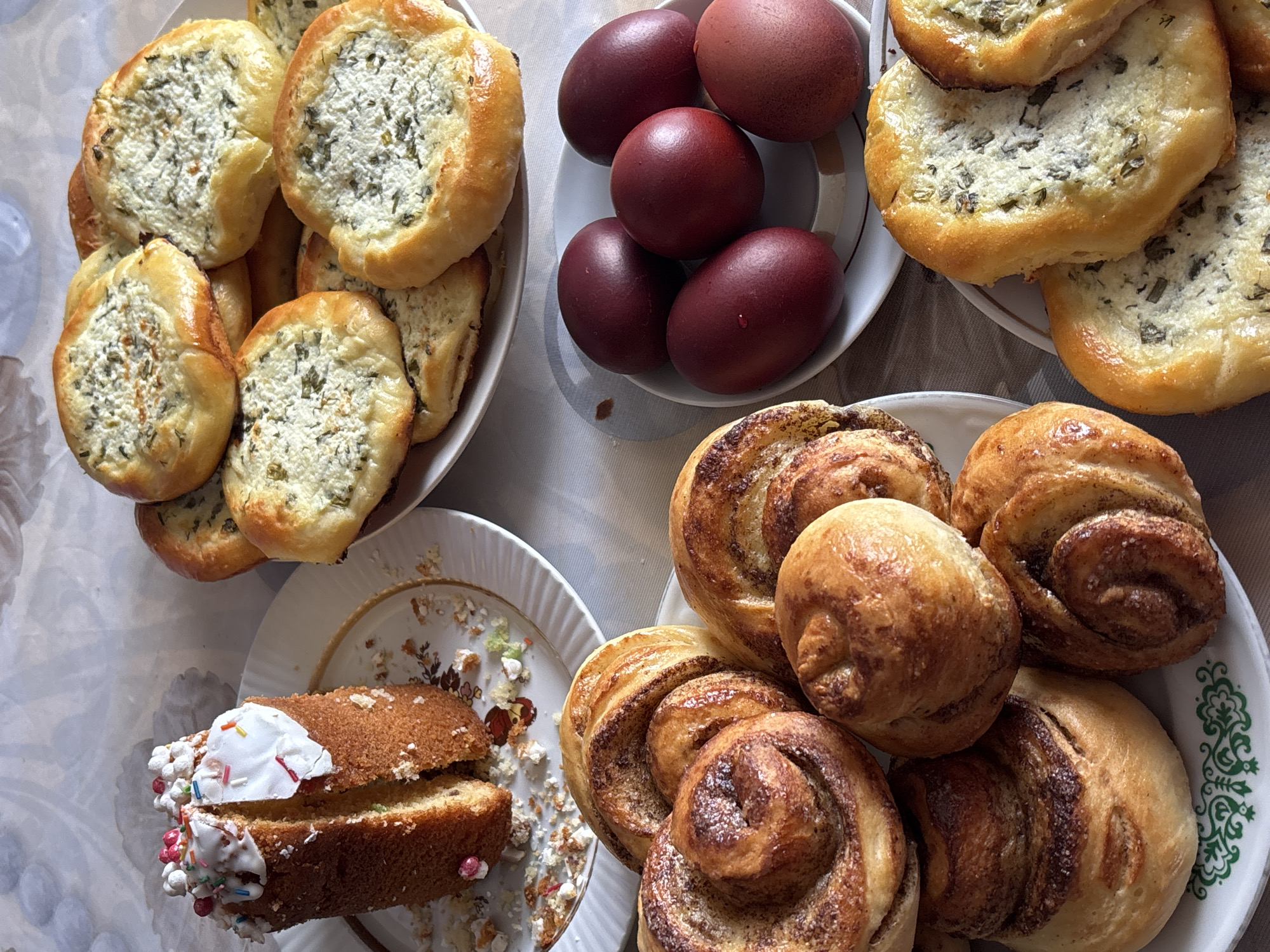The image shows a table with several plates of food. On the top left is a plate of cheese buns with herbs. Next to it is a plate with six red-brown colored eggs. On the top right is another plate with cheese buns with herbs. On the bottom right is a plate with cinnamon rolls. On the bottom left is a plate with a sliced cake with white icing and sprinkles. The plates are sitting on a white tablecloth. There is no indication of the city of Urada, Russia in the image.