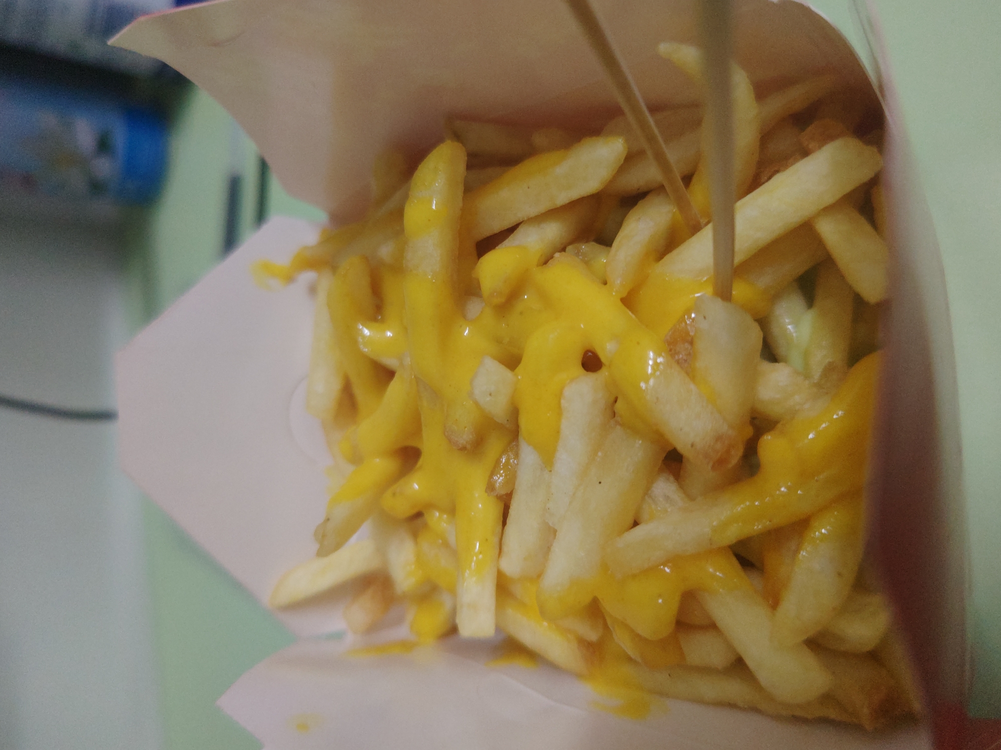 The image shows a serving of french fries covered in cheese sauce, contained within a white and tan paper container. A wooden skewer is present, piercing the fries. The photograph appears to be taken indoors with a blurred background suggesting an interior setting. The location is indicated as Chongqing High-tech Zone, China. No people are visible.
