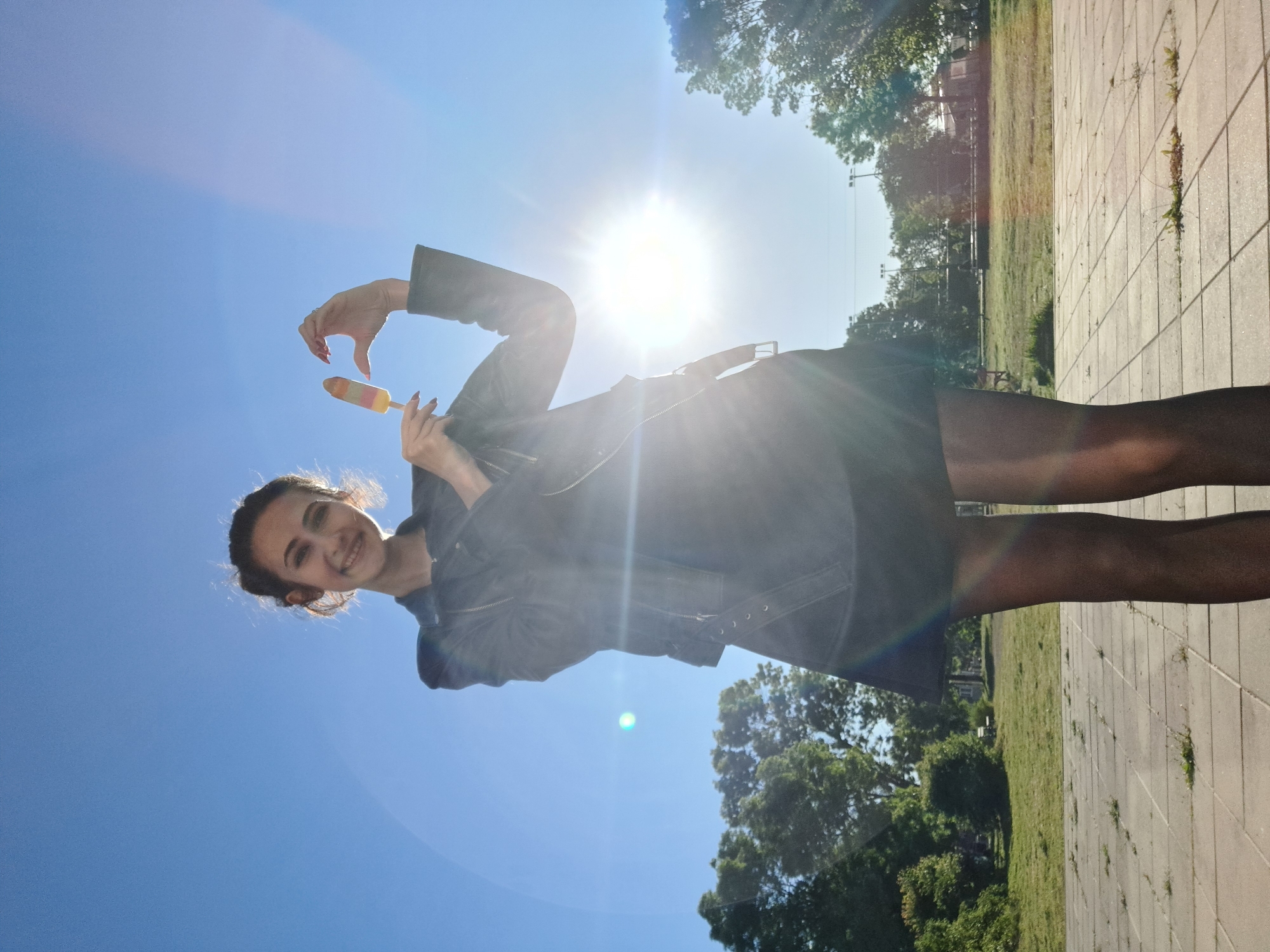 A young woman stands on a light gray concrete-tiled surface, holding a multi-colored ice pop up in her left hand, fingers curled towards the ice cream as if making a heart shape, while she holds the stick in her right hand. She smiles at the camera. She wears a black jacket and dark tights. A field of green grass and trees are visible on the right side of the image. The sky is bright blue with the sun directly overhead.