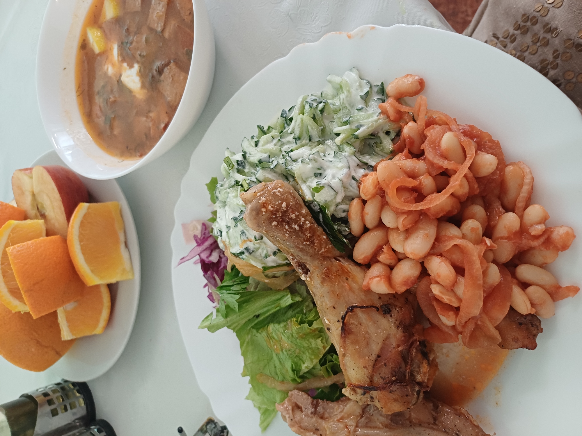 A white plate of food sits on a white tablecloth. The plate contains a portion of cooked chicken, a serving of white beans stewed with onions and tomato sauce, and a salad made of shredded cucumber and lettuce. A bowl of soup, with pieces of meat and vegetables, is to the right of the plate. In the background, a plate of sliced oranges and an apple sits on the table, along with a stainless steel utensil holder. The scene appears to be set in a restaurant in the city of Varna, Bulgaria, as suggested by the style of the tableware and the overall presentation, which is typical of Bulgarian cuisine. There are no people visible in the image.