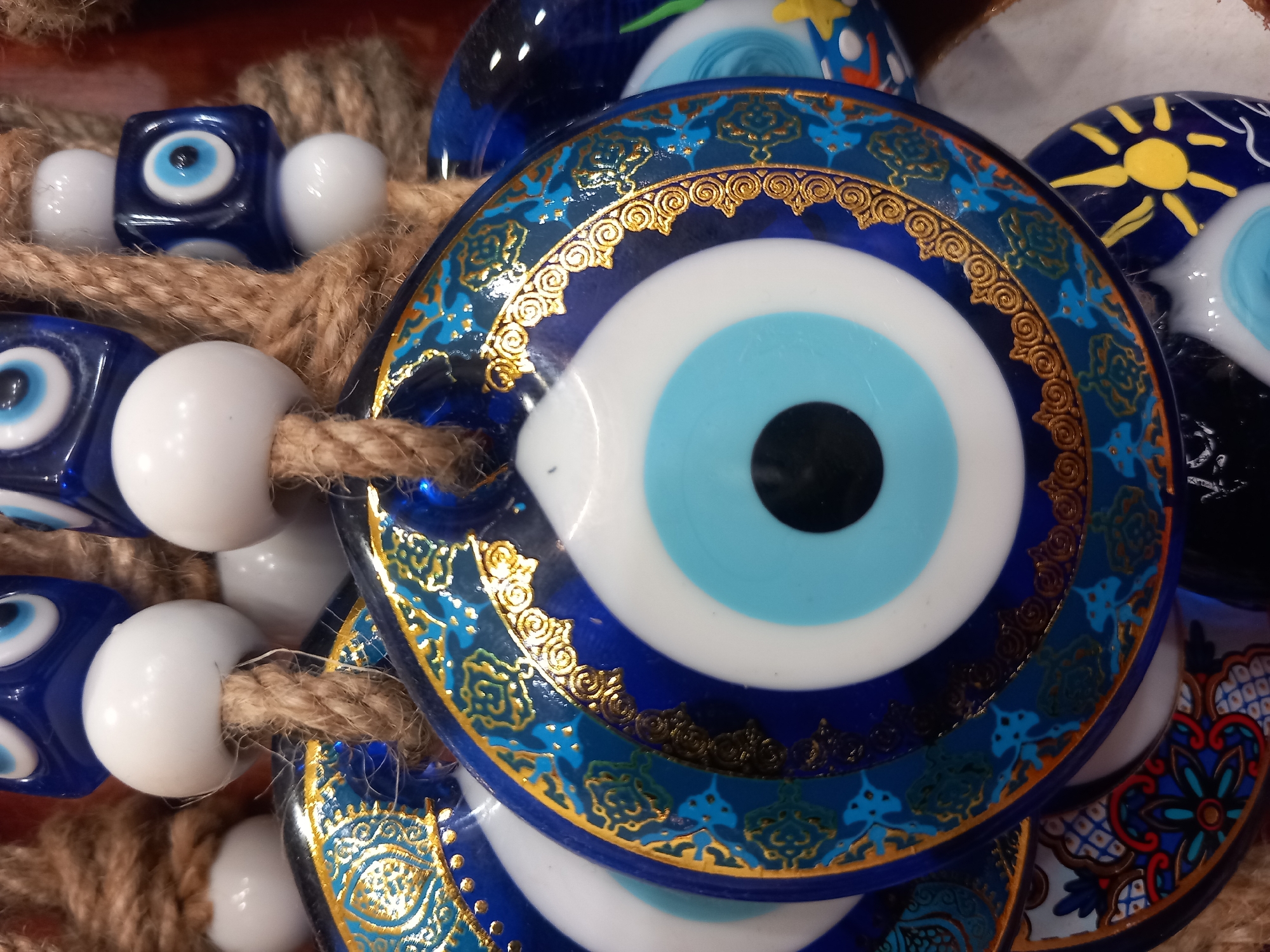 A close-up, eye-level shot showcases a collection of traditional evil eye charms, likely from Kos, Greece, hanging from a rough, brown twine. The charms are predominantly blue and white, featuring the iconic concentric circles of an eye. Some are round glass pendants adorned with intricate gold detailing and blue geometric patterns. Others are smaller, cube-shaped beads, also depicting the evil eye motif. White spherical beads are interspersed among the charms on the twine. The background is softly blurred, hinting at a display or market setting, with warm reddish-brown tones and other colors possibly visible. The focus is sharp on the central charms, highlighting their textures and decorative elements.